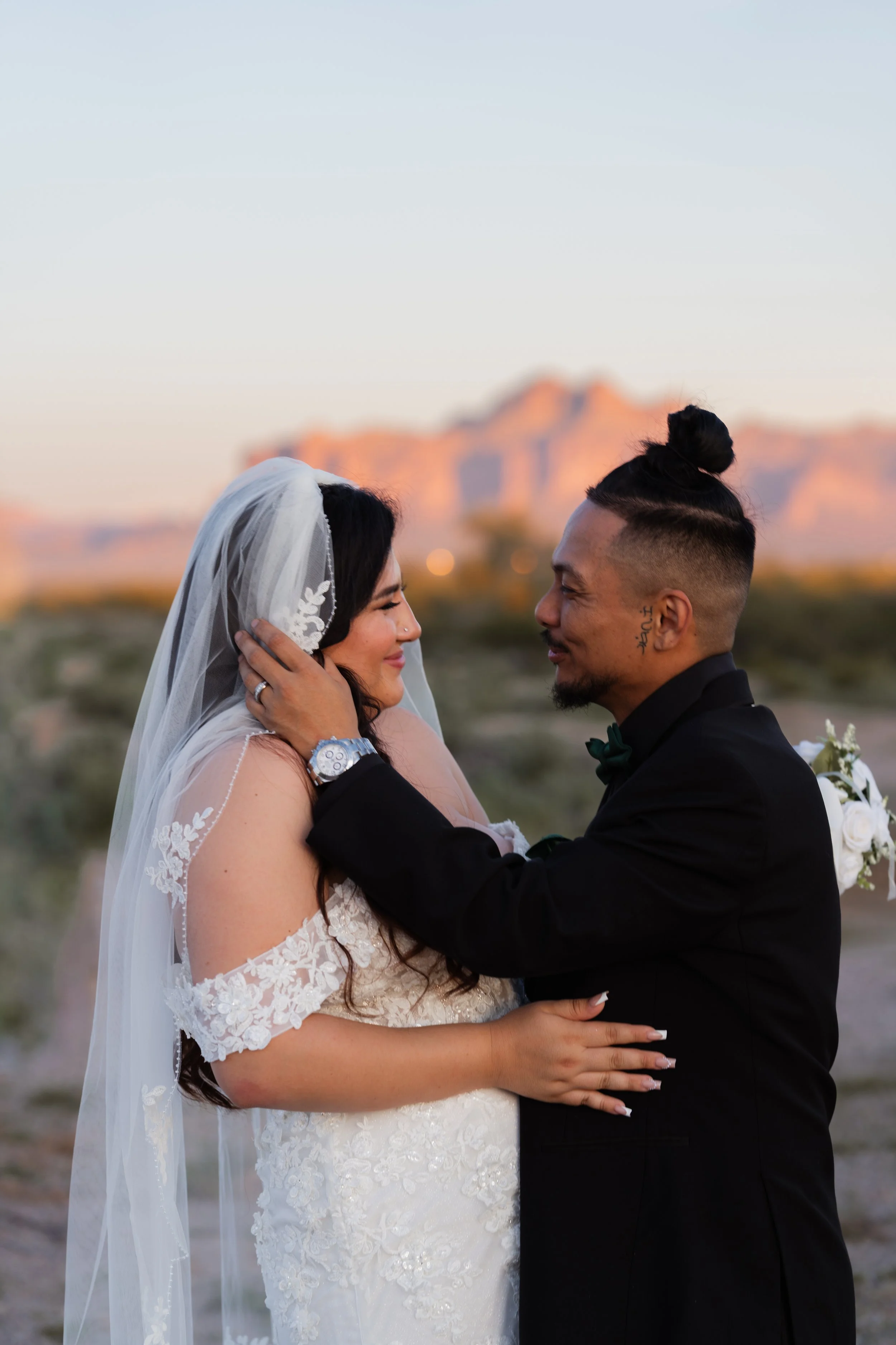 A bride and groom sharing an intimate moment outdoors during sunset, with desert landscape and mountains in the background.