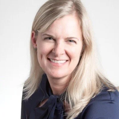 A woman with long blonde hair smiling at the camera, wearing a navy blouse with a bow.