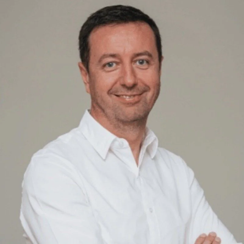 Portrait of a man with short dark hair, wearing a white button-up shirt, smiling, against a plain background.