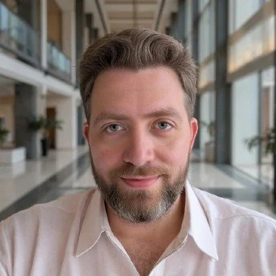 A man with light skin, brown hair, and a beard taking a selfie indoors, with large windows and a modern building interior behind him.