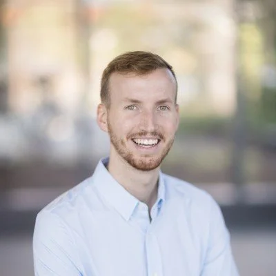 A smiling man with short hair and a beard, wearing a light blue button-up shirt outdoors with a blurred background of trees and buildings.