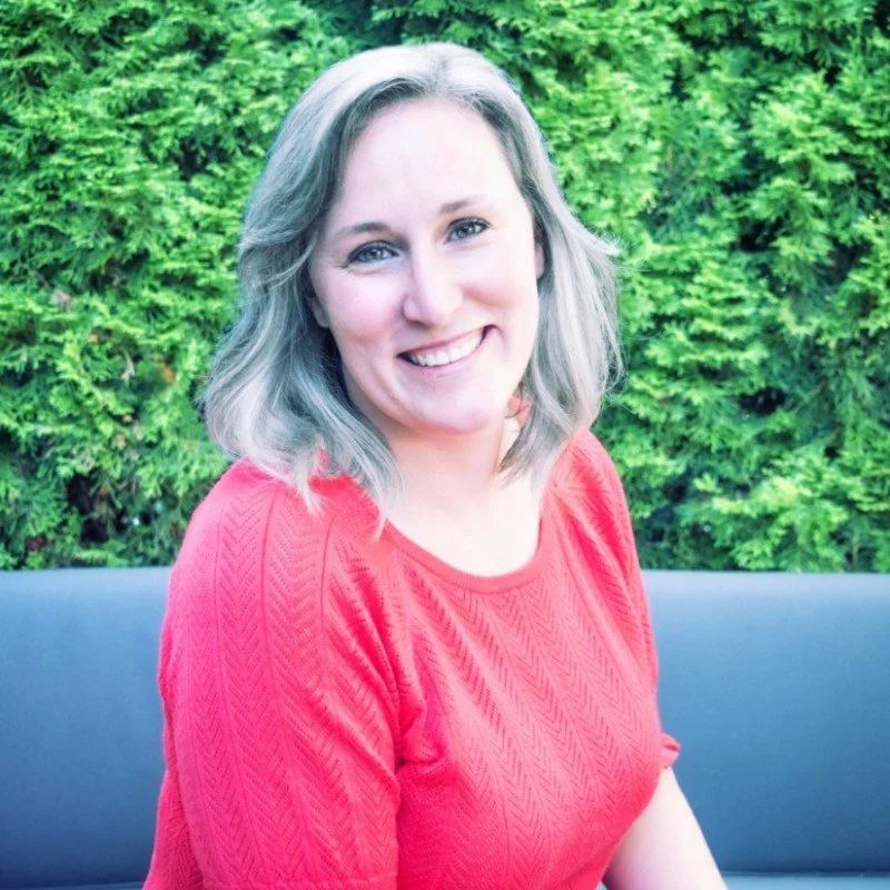 A smiling woman with shoulder-length gray hair, wearing a red sweater, sitting outdoors in front of green foliage.