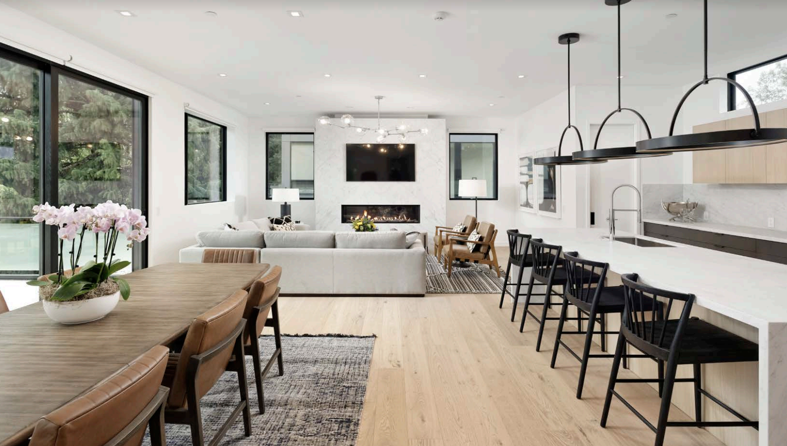 Modern open-concept living room and kitchen with white walls, large windows, light wood flooring, a dining table with chairs, a kitchen island with bar stools, and a cozy seating area with sofas and armchairs