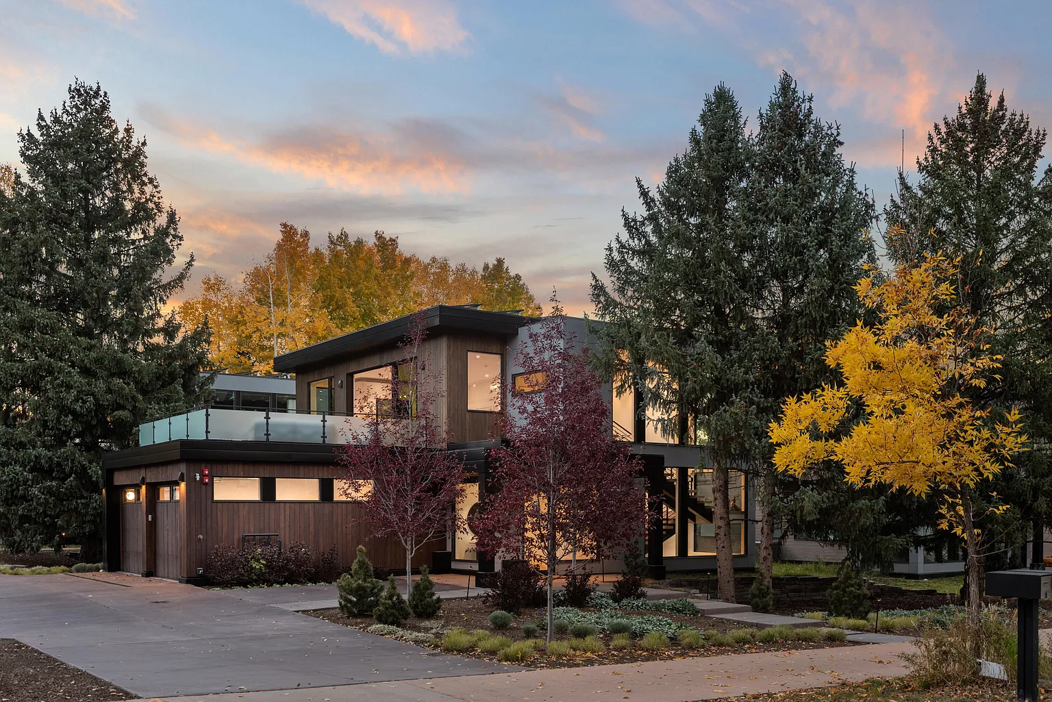 Modern two-story house with wooden exterior, large windows, surrounded by colorful trees, during sunset.