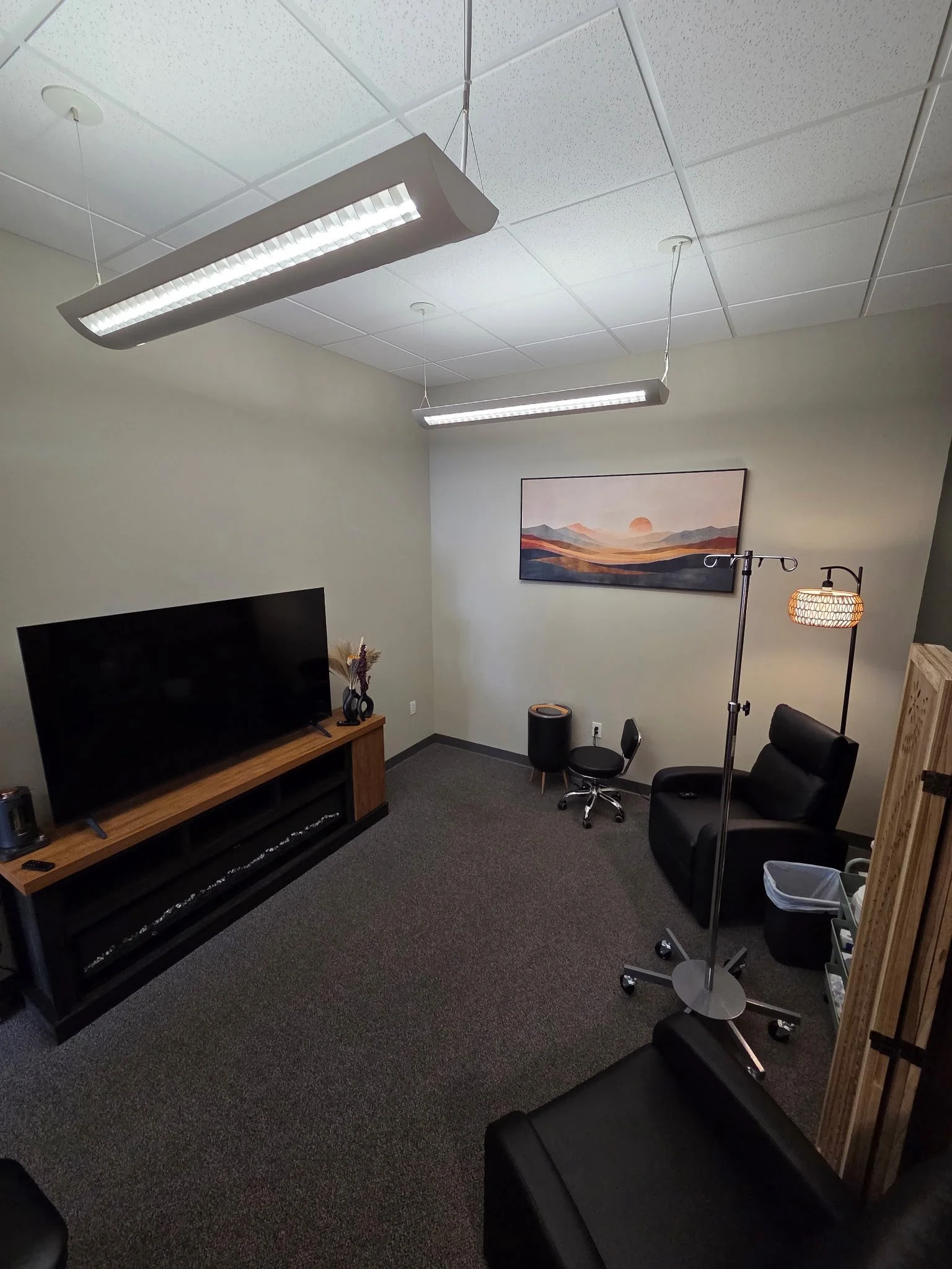 A small, sparsely furnished office or therapy room with a television, two ceiling lights, a painting on the wall illustrates a sunset over mountains, a black leather chair, a small armchair, a standing lamp, a rolling chair, a coat rack or IV pole, a small table, and some storage bins.