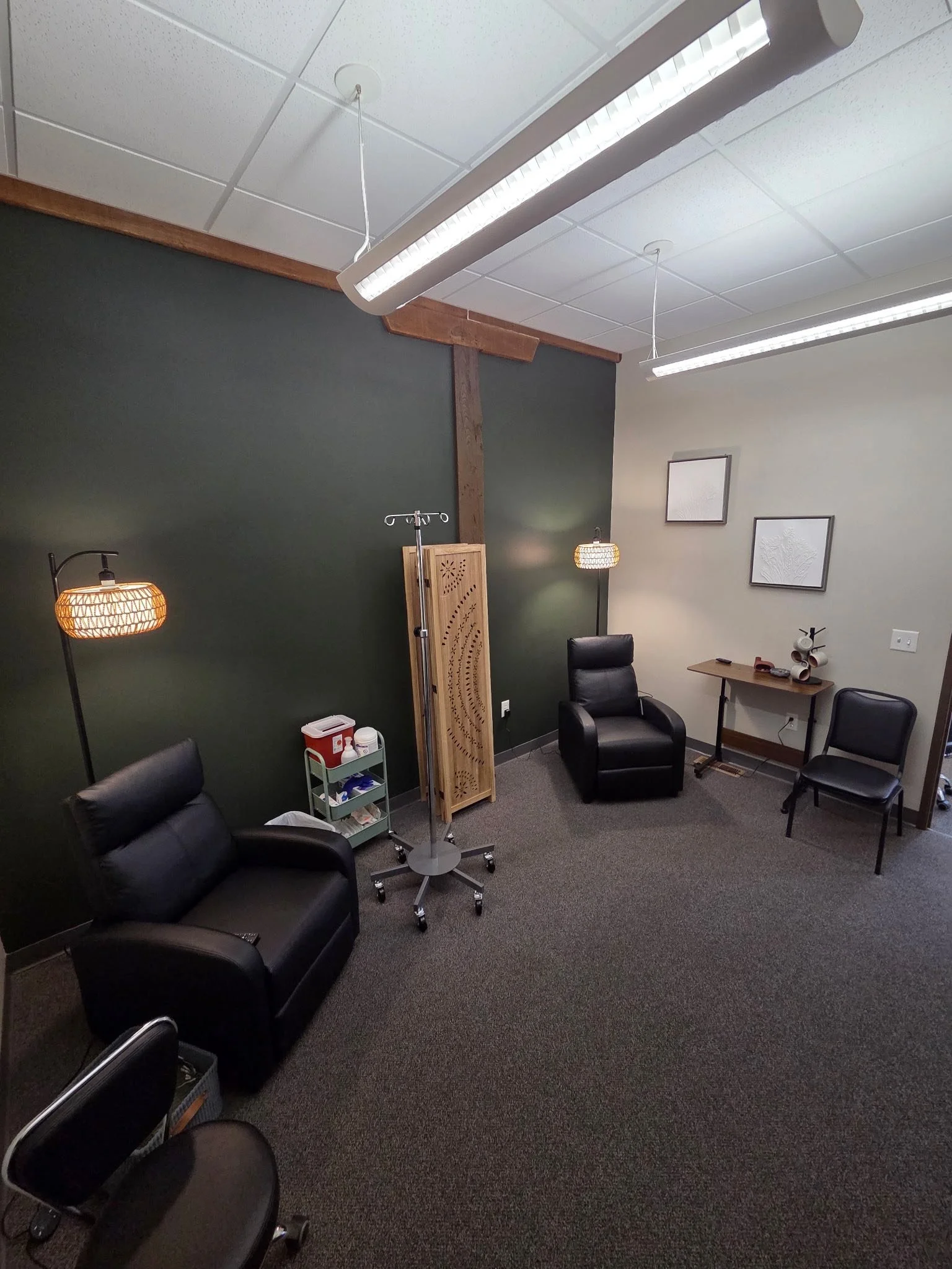 A small medical or therapy room with two black leather chairs, a rolling stool, and a small table with medical supplies. The room has a dark green accent wall, framed artwork, floor lamps, and overhead fluorescent lighting.