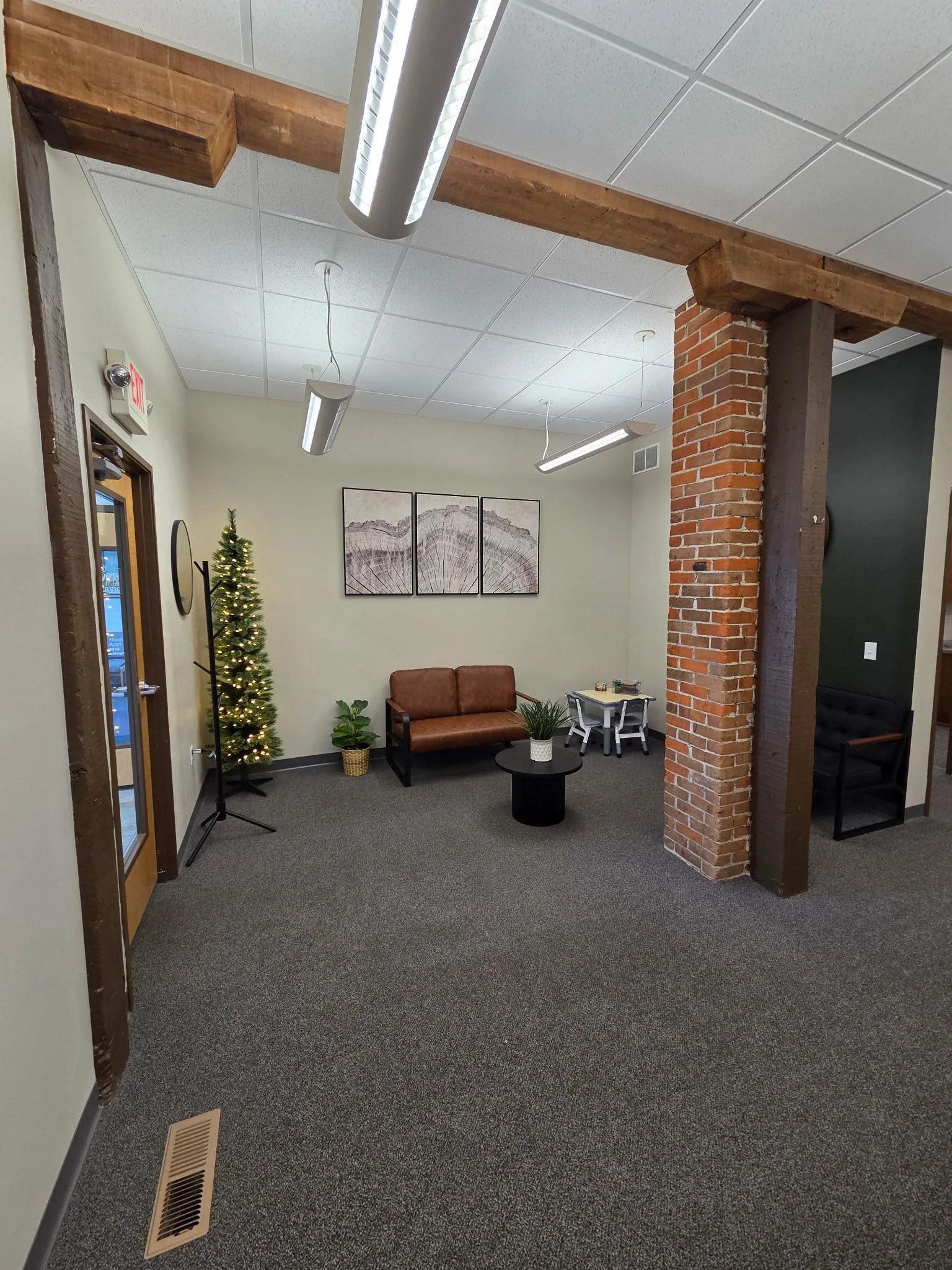 A cozy waiting room with a small Christmas tree, a brown leather loveseat, framed artwork on the wall, and a small table with a potted plant. There are wooden beams, exposed brick, and modern lighting fixtures ceiling.