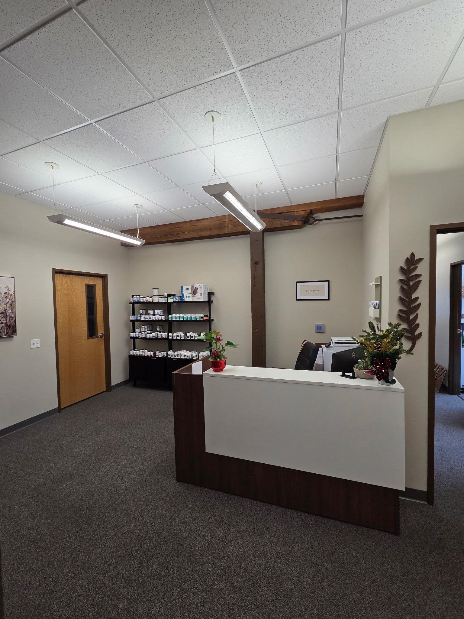 Reception area with a white desk, potted plant, black chair, and shelves with bottles, in a professional office space.