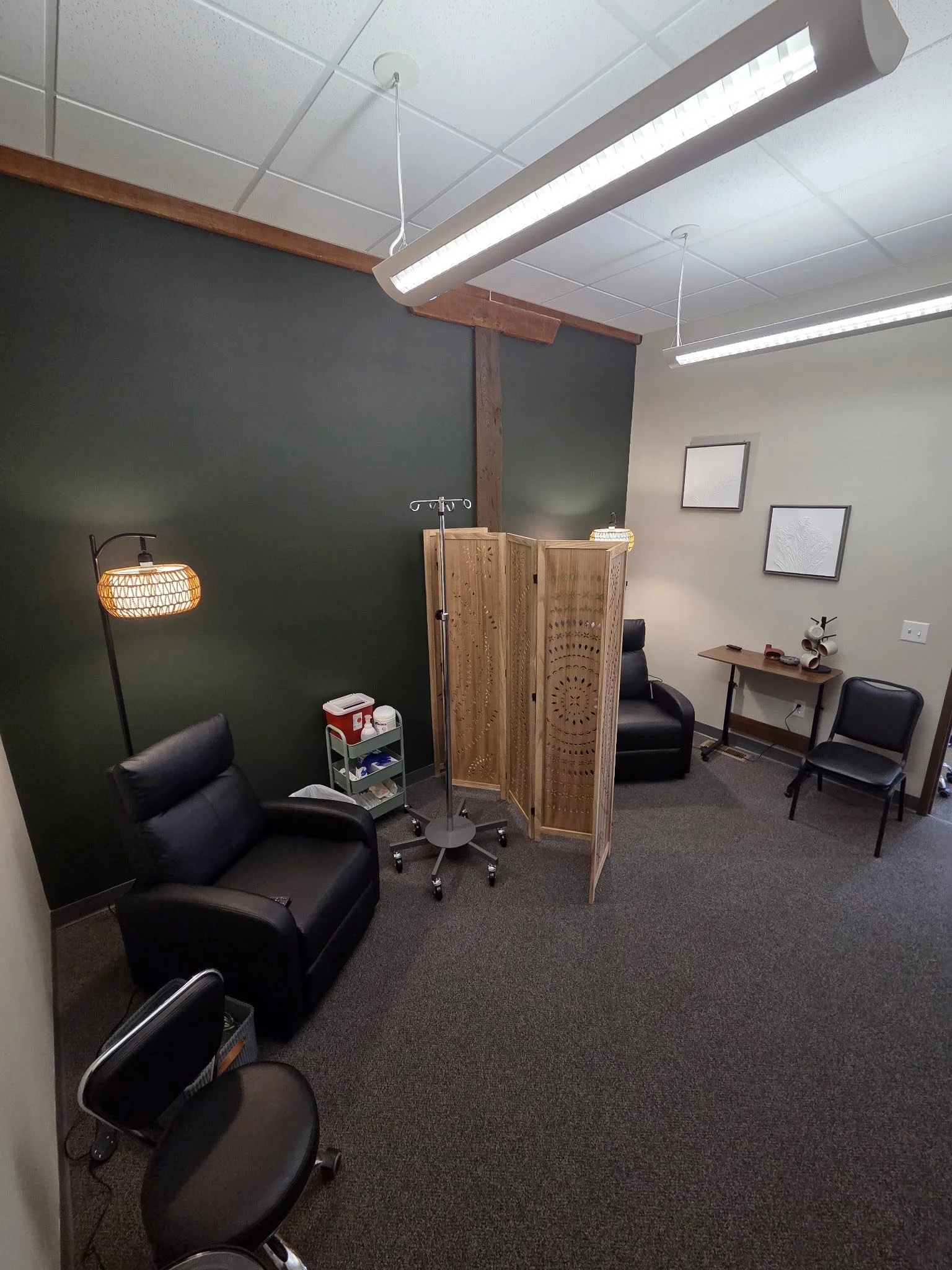 A small medical or therapy office with two black leather chairs, a wooden privacy divider, a medical IV stand, a small cart with supplies, and a desk with two photos on the wall.