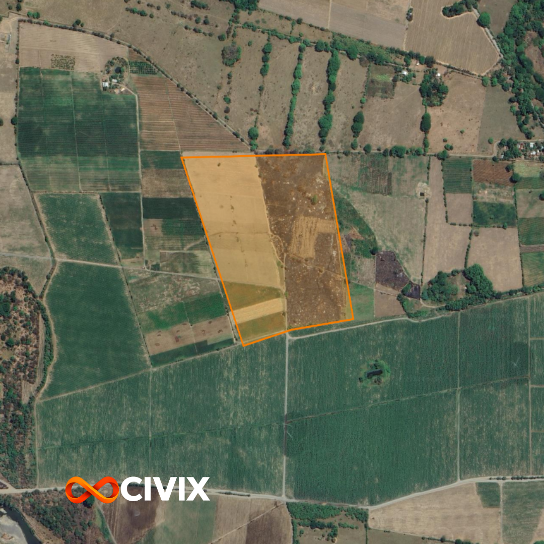 Foto aérea de un terreno agrícola delimitado con línea naranja, rodeado de diferentes campos de cultivo y áreas con árboles.