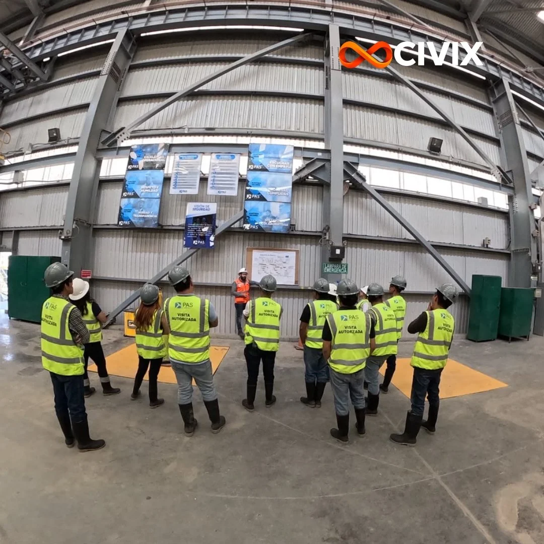 Grupo de personas con chalecos y cascos de seguridad en una visita guiada a una fábrica o planta industrial, frente a una estructura metálica grande con carteles y señalización.