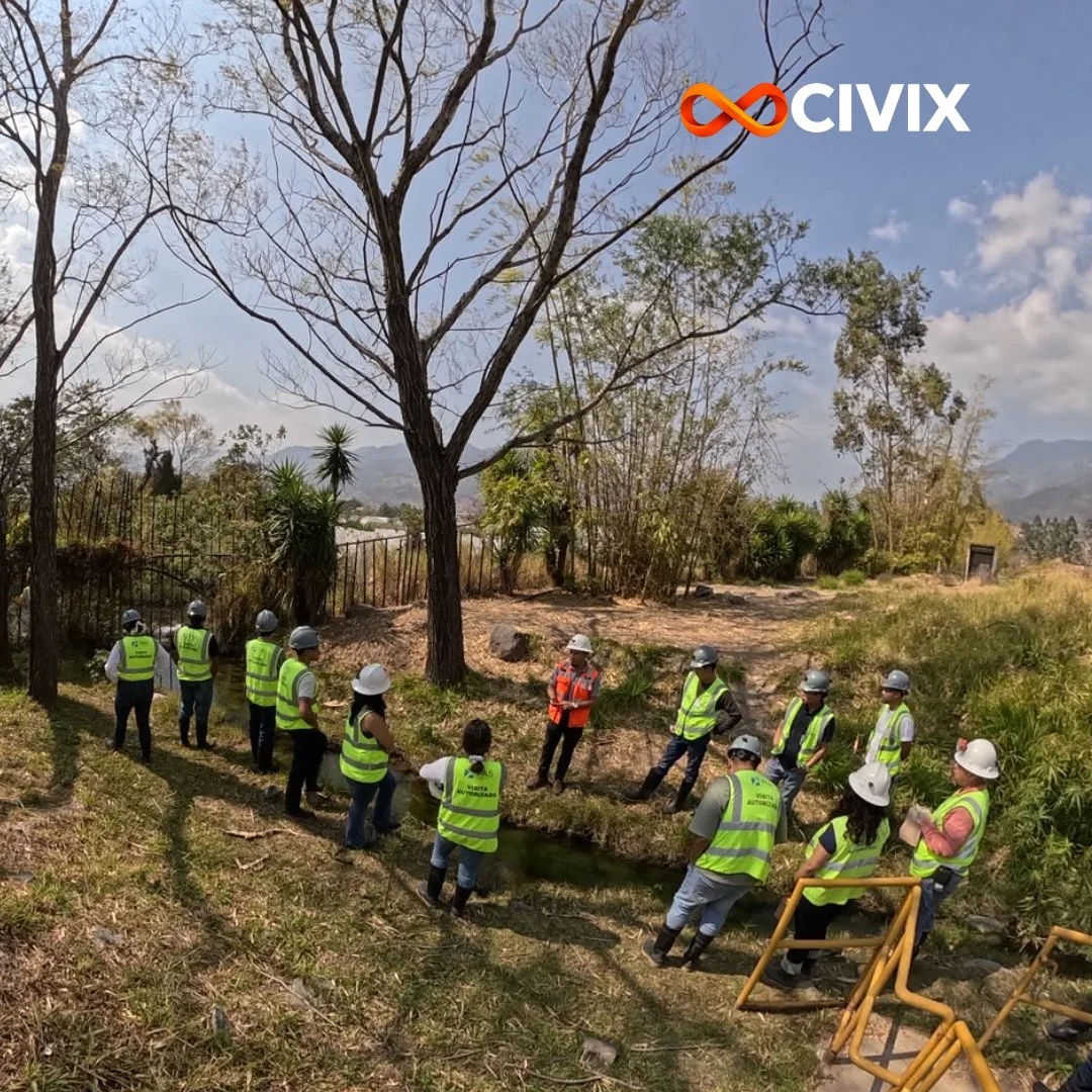 Grupo de personas con cascos y chalecos amarillos observan un río en un área natural, con árboles y montañas al fondo durante el día.