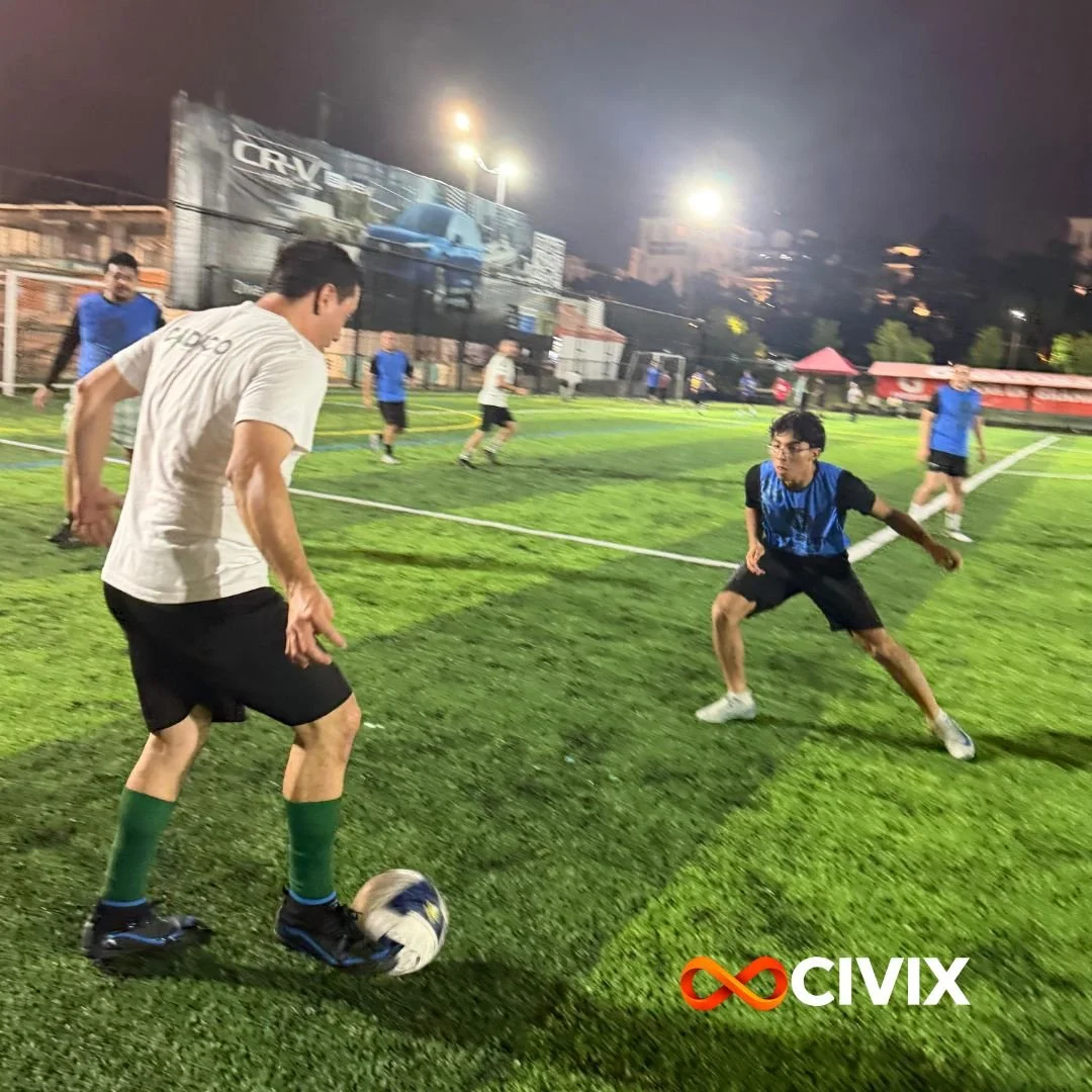 Grupo de jóvenes jugando fútbol en un campo de césped artificial por la noche, con iluminación y publicidad en el fondo, y el logo de Civix en la esquina inferior derecha.