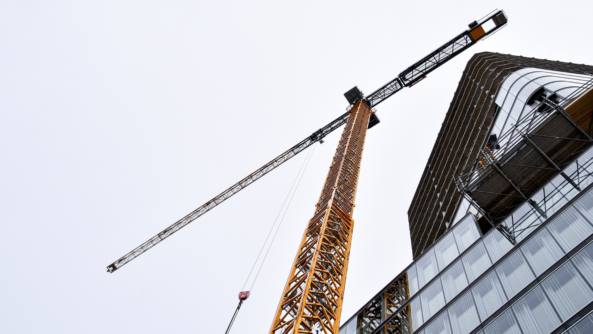 Construcción de un edificio moderno con fachada de vidrio y un enredado de estructuras metálicas, junto a una torre de grúa de gran altura con una pluma larga y un contrapeso en la parte superior.