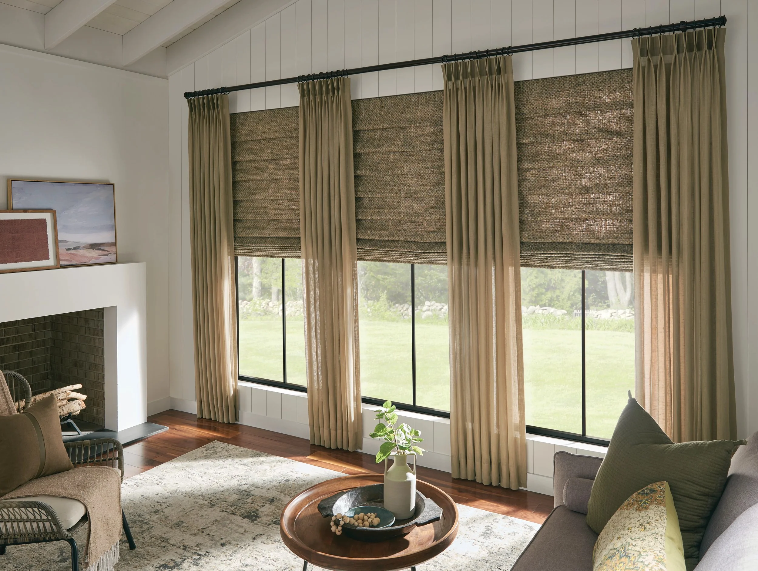 A cozy living room with large windows covered by brown and beige curtains, a fireplace, a light-colored area rug, a coffee table, and comfortable chairs.
