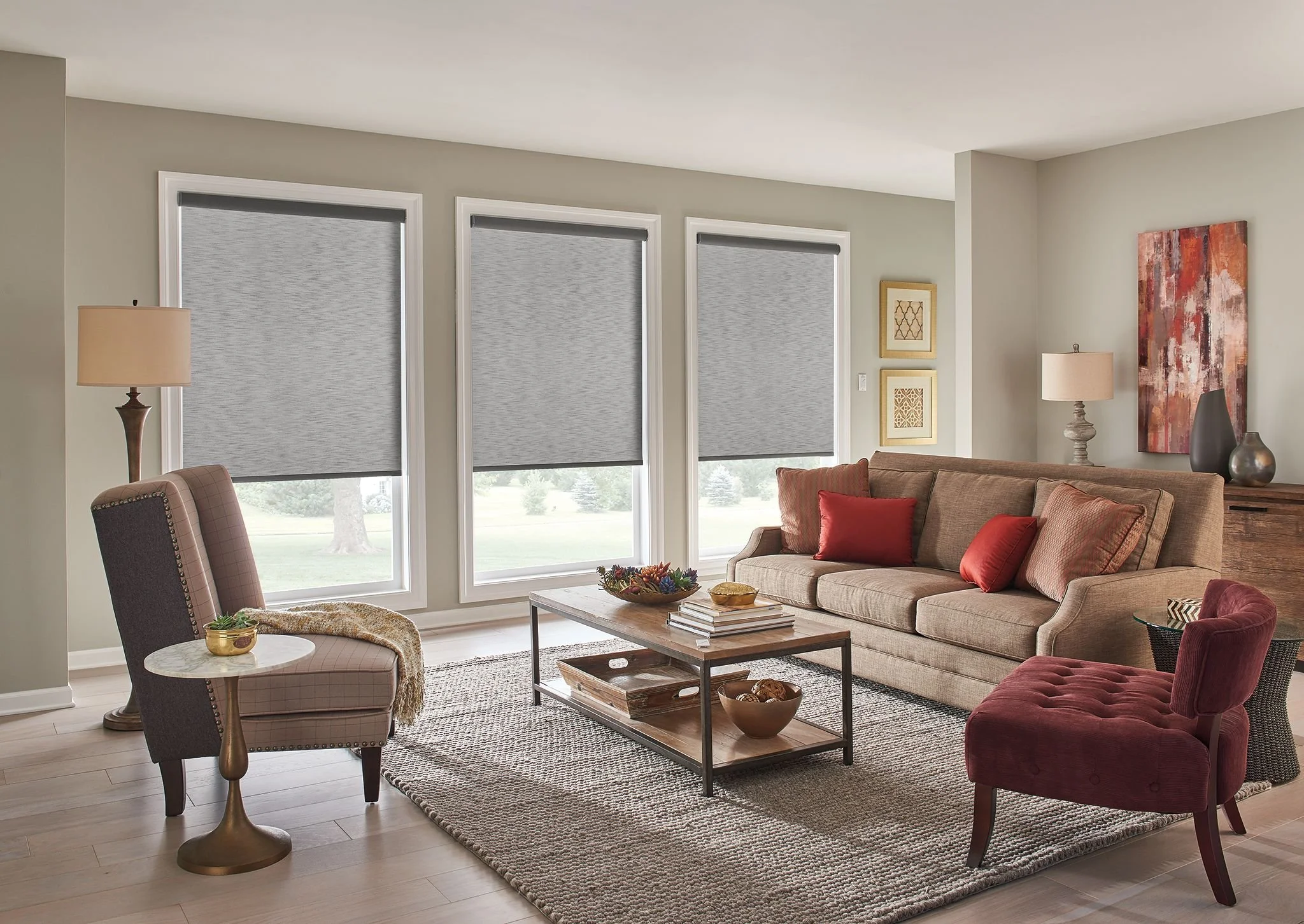 Living room with beige sofa, red pillows, wooden coffee table, patterned rug, beige armchair, side table with a gold vase, table lamp, large windows with gray blinds, walls with framed artwork, and wooden flooring.