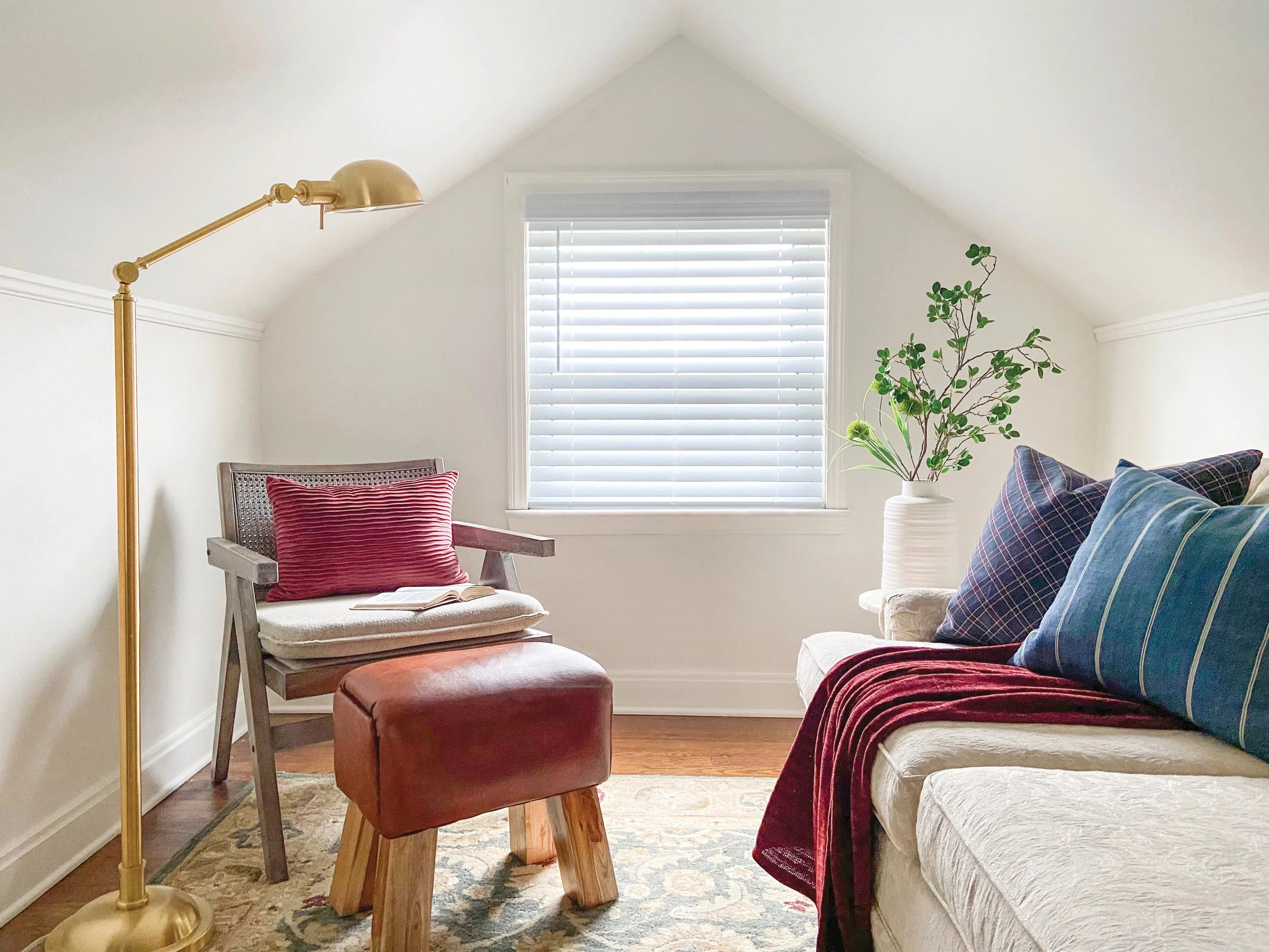 A cozy living room with a white sofa adorned with blue and red checkered pillows, a white lamp with a brass arm, a wooden chair with a red striped pillow, and a small leather ottoman on a patterned rug. There is a large window with white blinds, and a leafy green plant in a white vase on a side table.