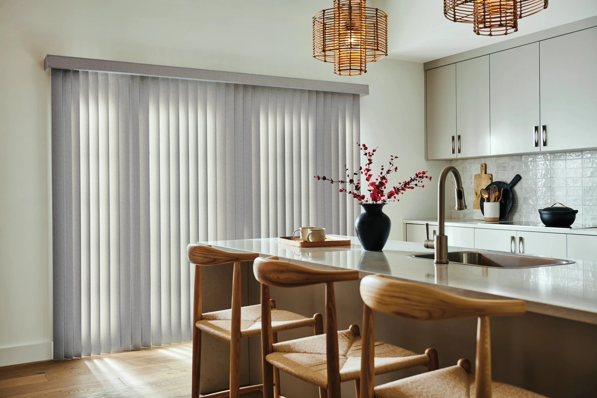 Kitchen with beige cabinets, a white countertop, a black vase with red flowers, wooden chairs, and gray vertical blinds