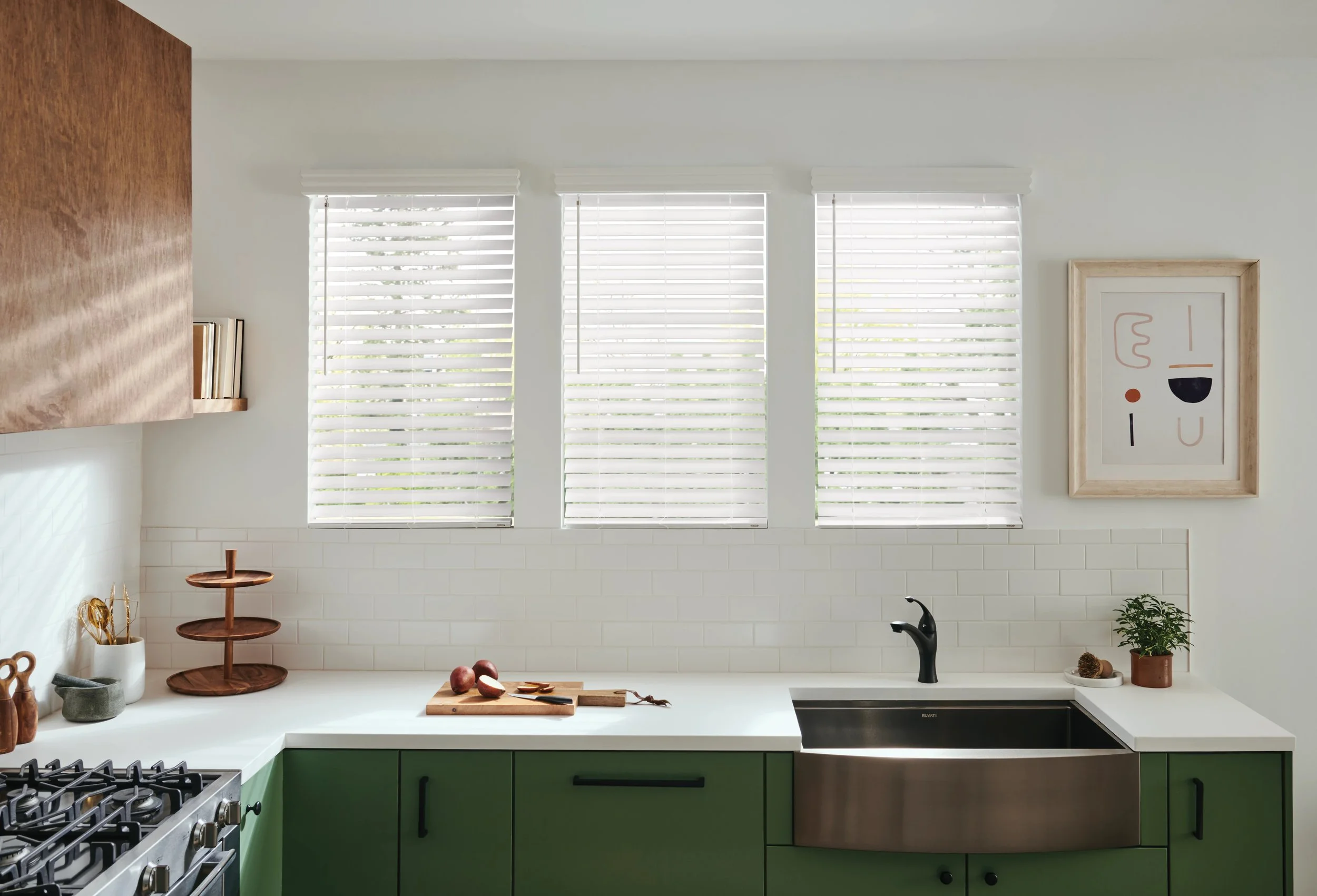 Modern kitchen with three large windows covered with white blinds, a white countertop, green cabinets, a black sink, and minimalist decor including a framed abstract art piece and wooden trays with onions.