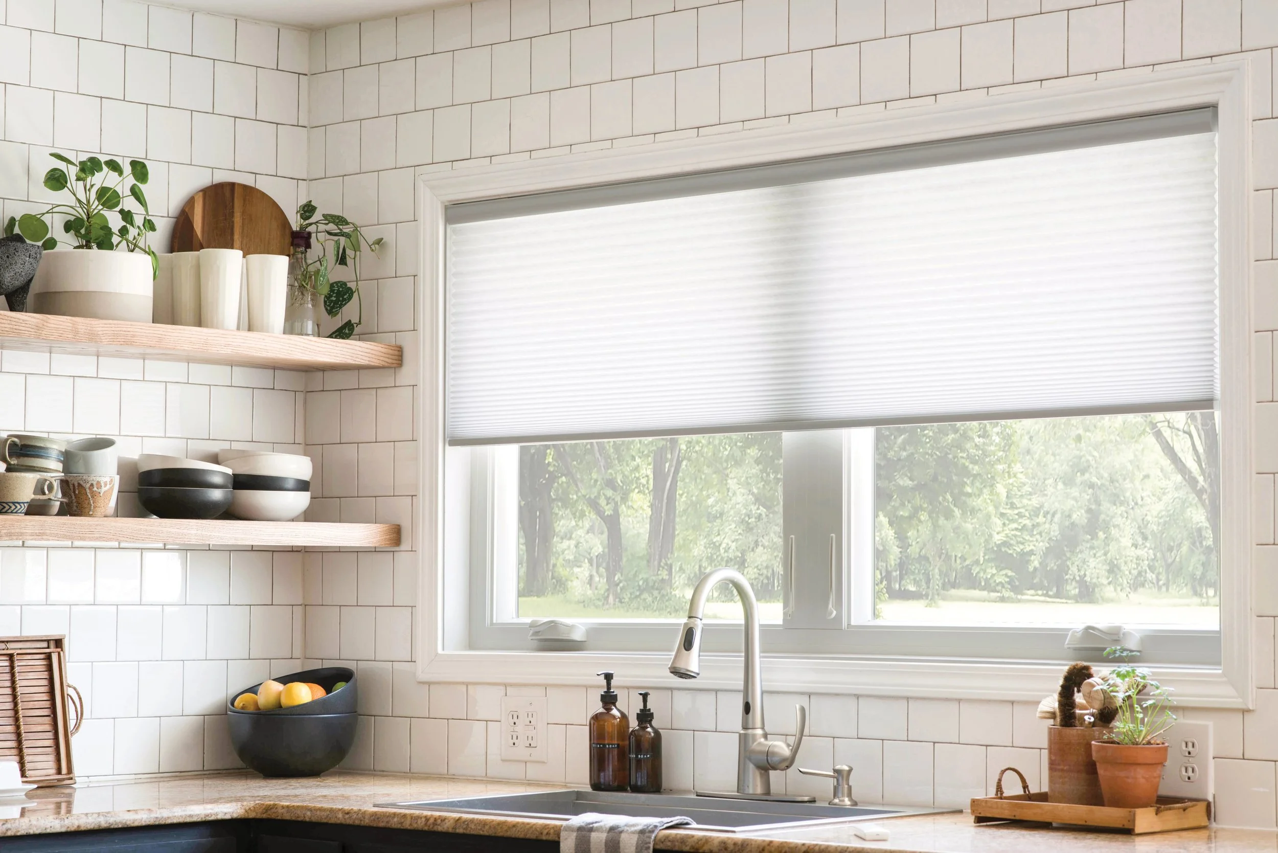 White kitchen with a window and white tiled walls. Wooden shelves holding plants and kitchenware. Counter with fruit bowl, soap dispensers, plants, and a kitchen towel.