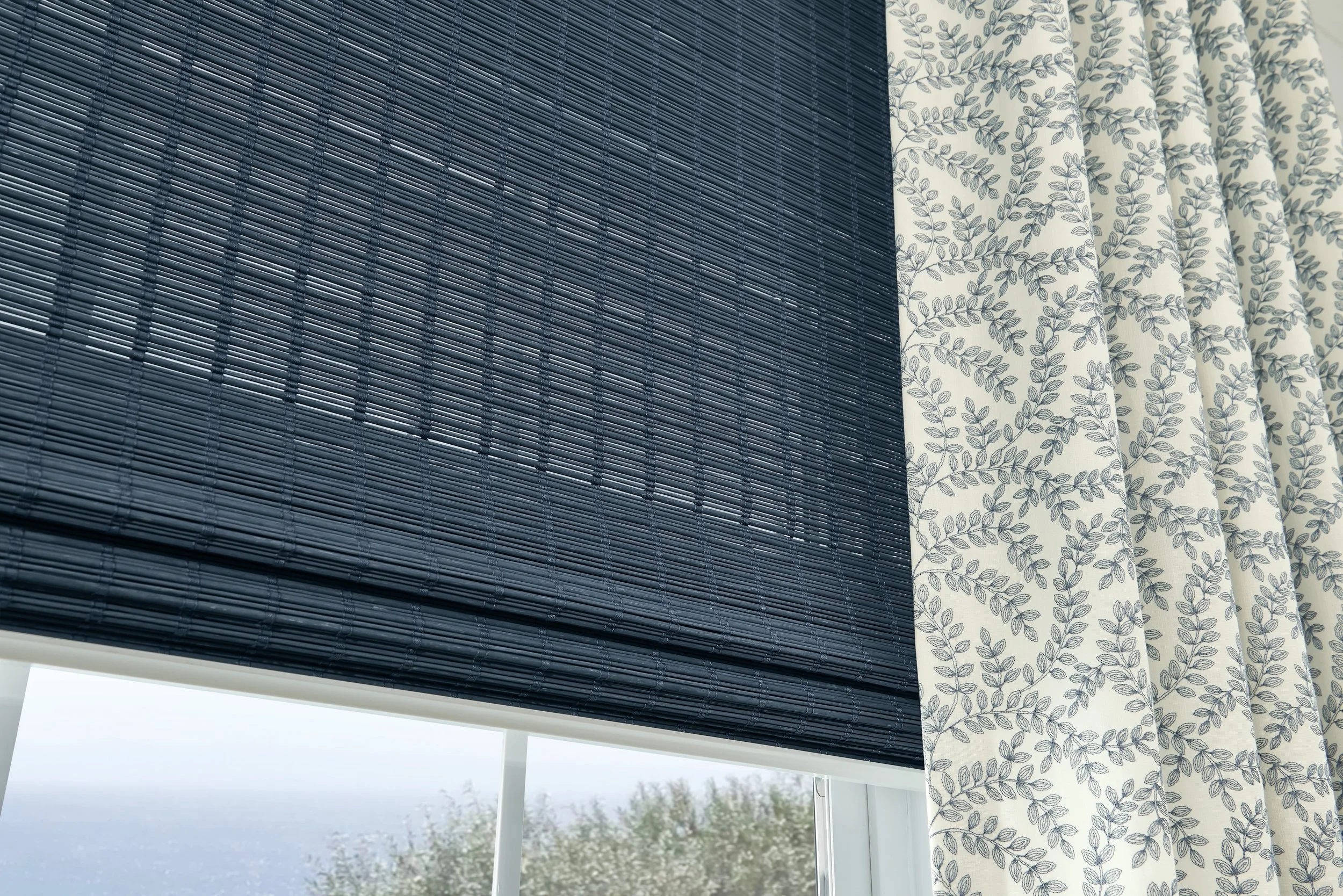 A view of a window with a dark blue woven bamboo shade and a white curtain with a blue leaf pattern, with a cloudy sky and some trees visible outside.