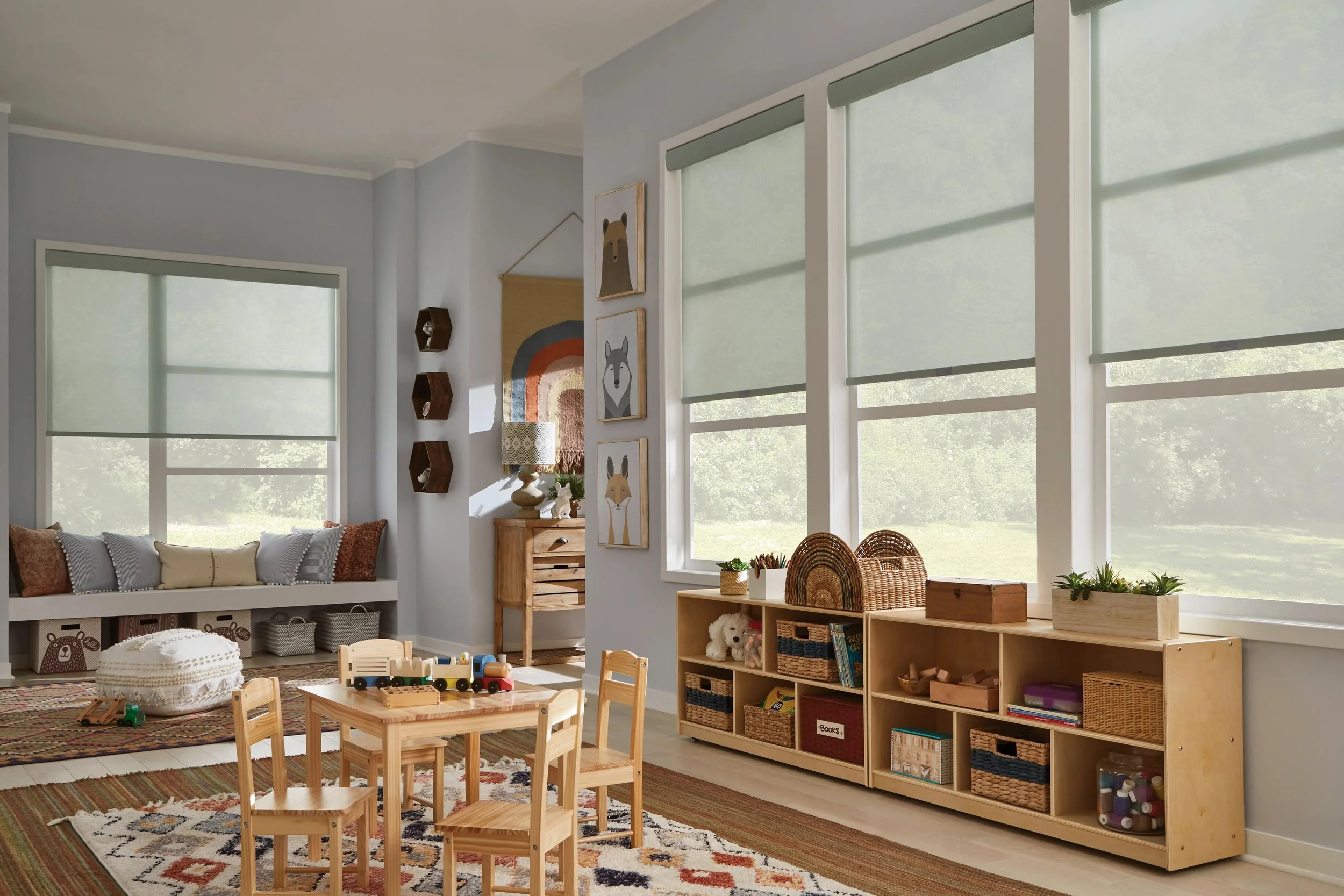A cozy children's playroom with large windows covered by light green shades, a built-in window seat with colorful throw pillows, wall art featuring animal illustrations, a wooden dollhouse, a toy train on a small table, woven baskets, a plush teddy bear, and shelves filled with books and toys, all on a multicolored rug.