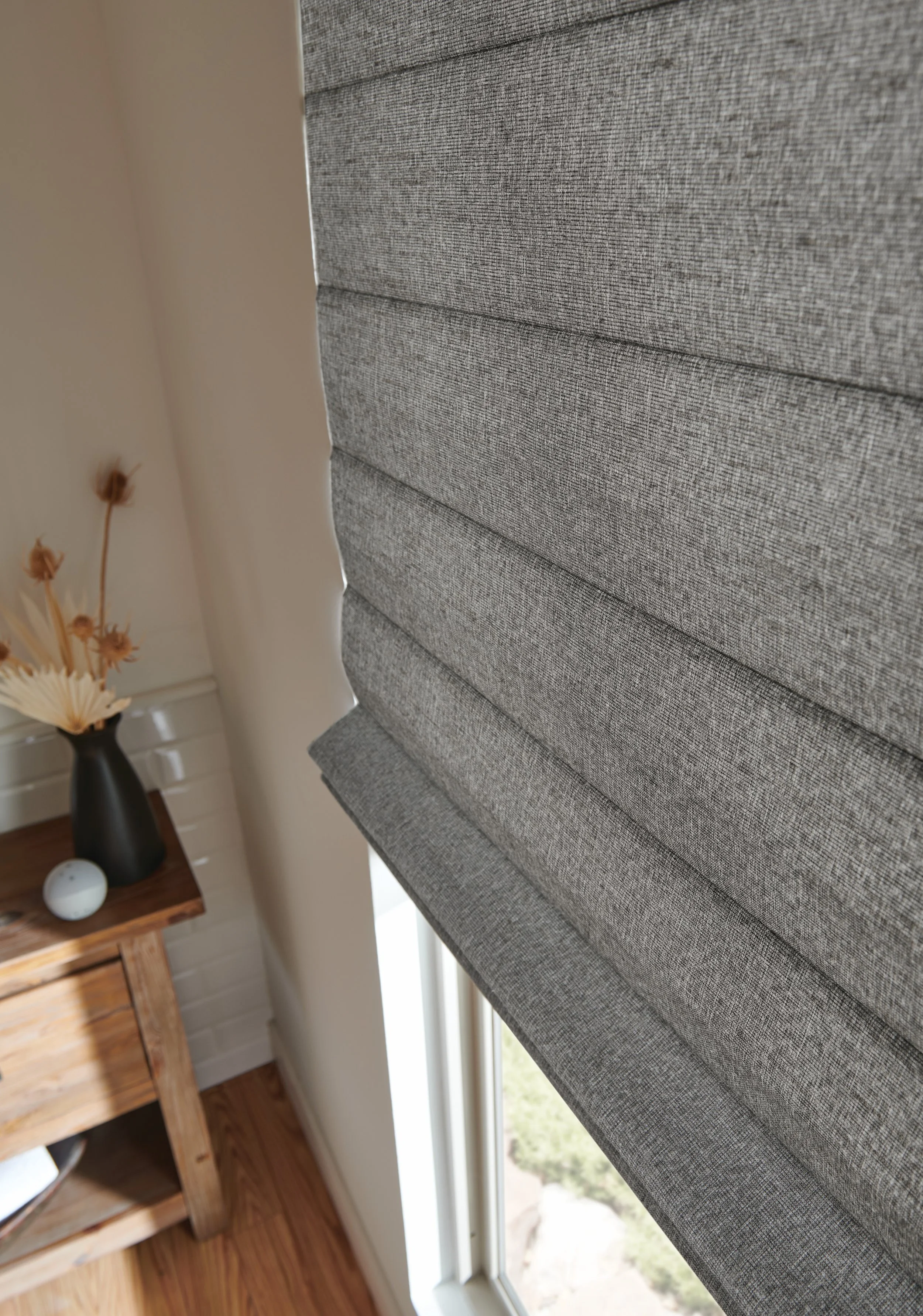 Close-up of a textured gray upholstered headboard in a bedroom, with part of a window and outside view visible to the right, and a wooden table with decorative items in the background.