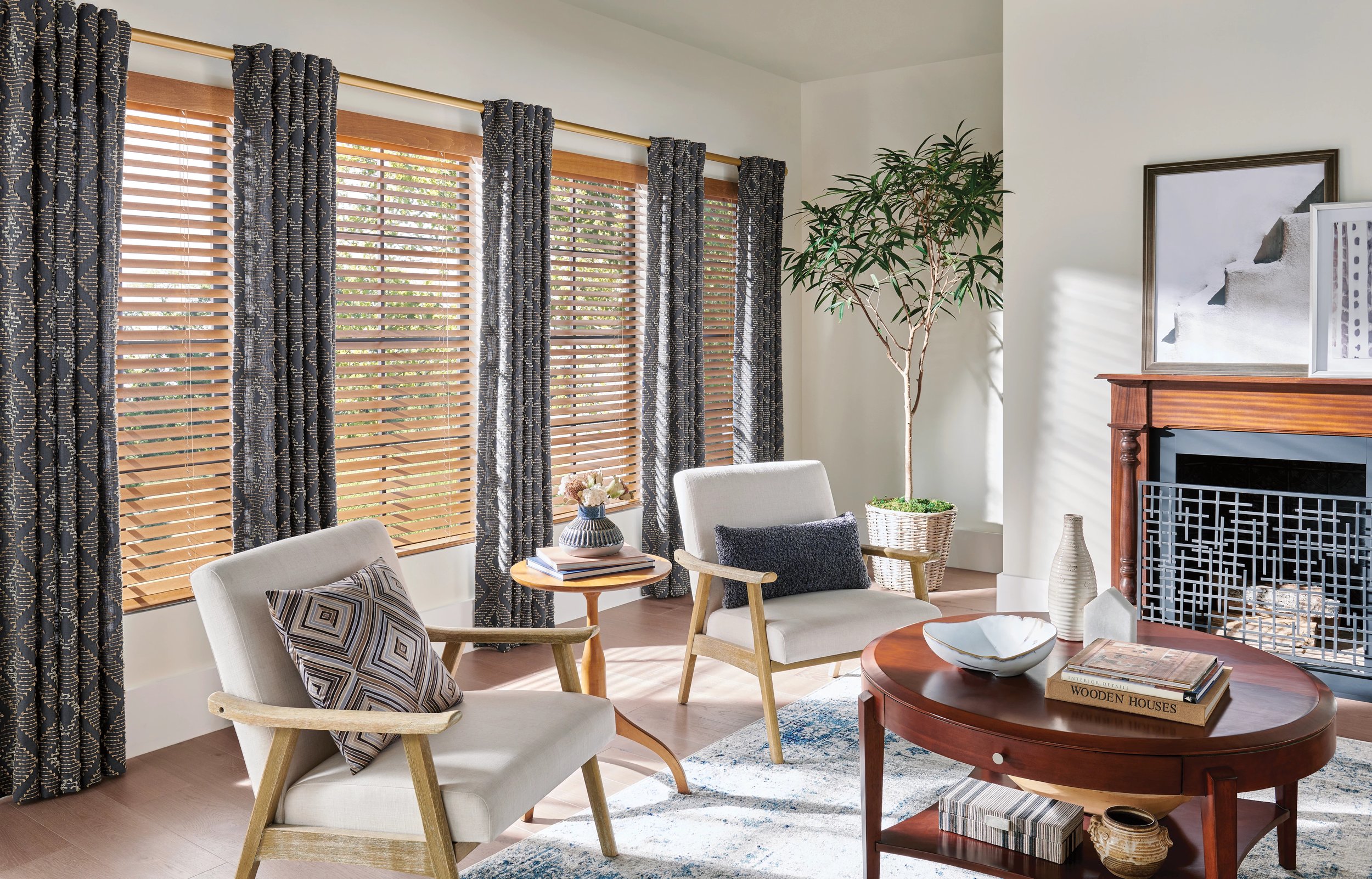 Living room with beige armchairs, a round wooden coffee table, a fireplace, and large windows with wooden blinds and patterned curtains, decorated with plants and artwork.