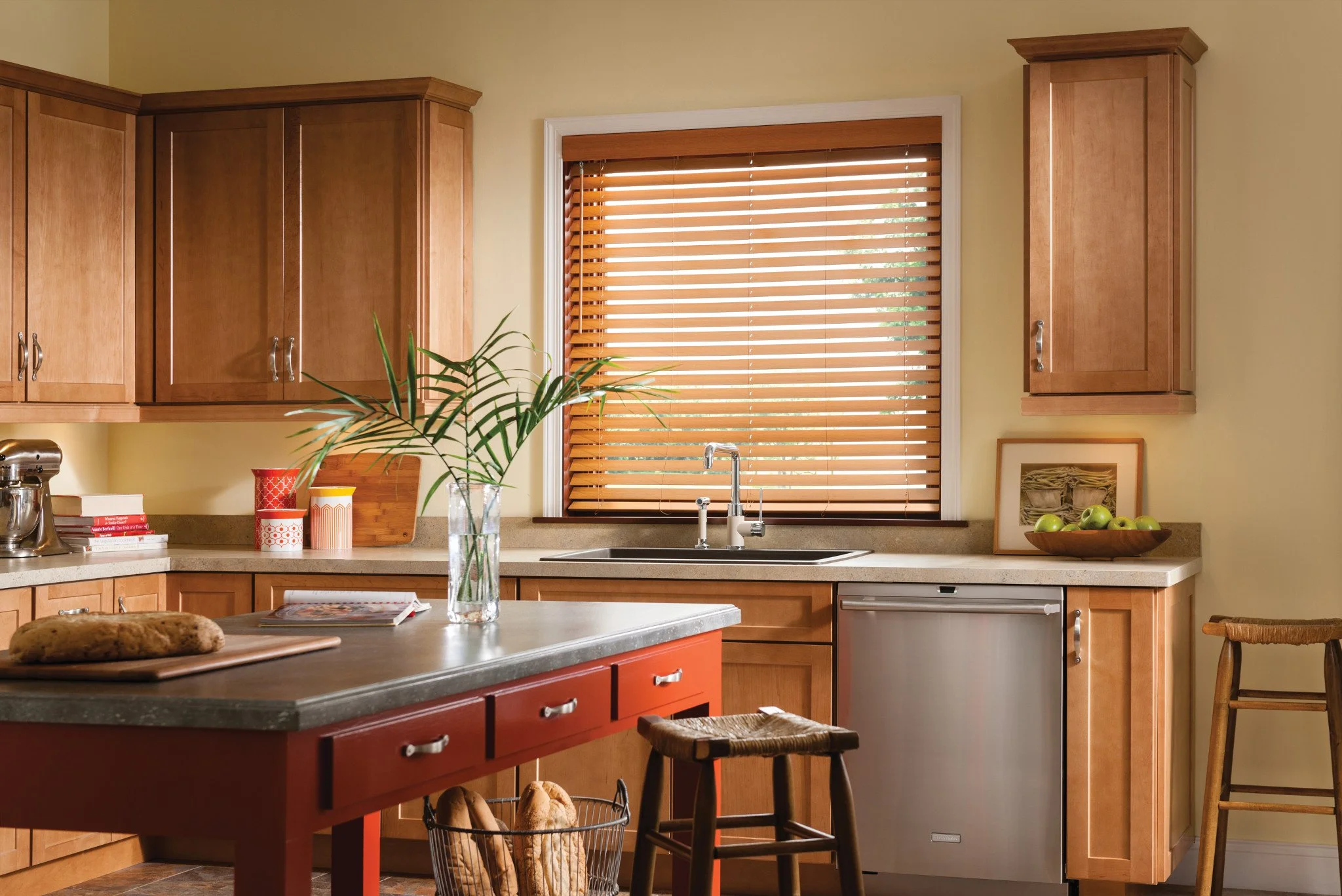 A kitchen with wooden cabinets, a window with wooden blinds, a granite countertop, and various kitchen items including a plant in a glass vase, a bowl of green apples, and books.