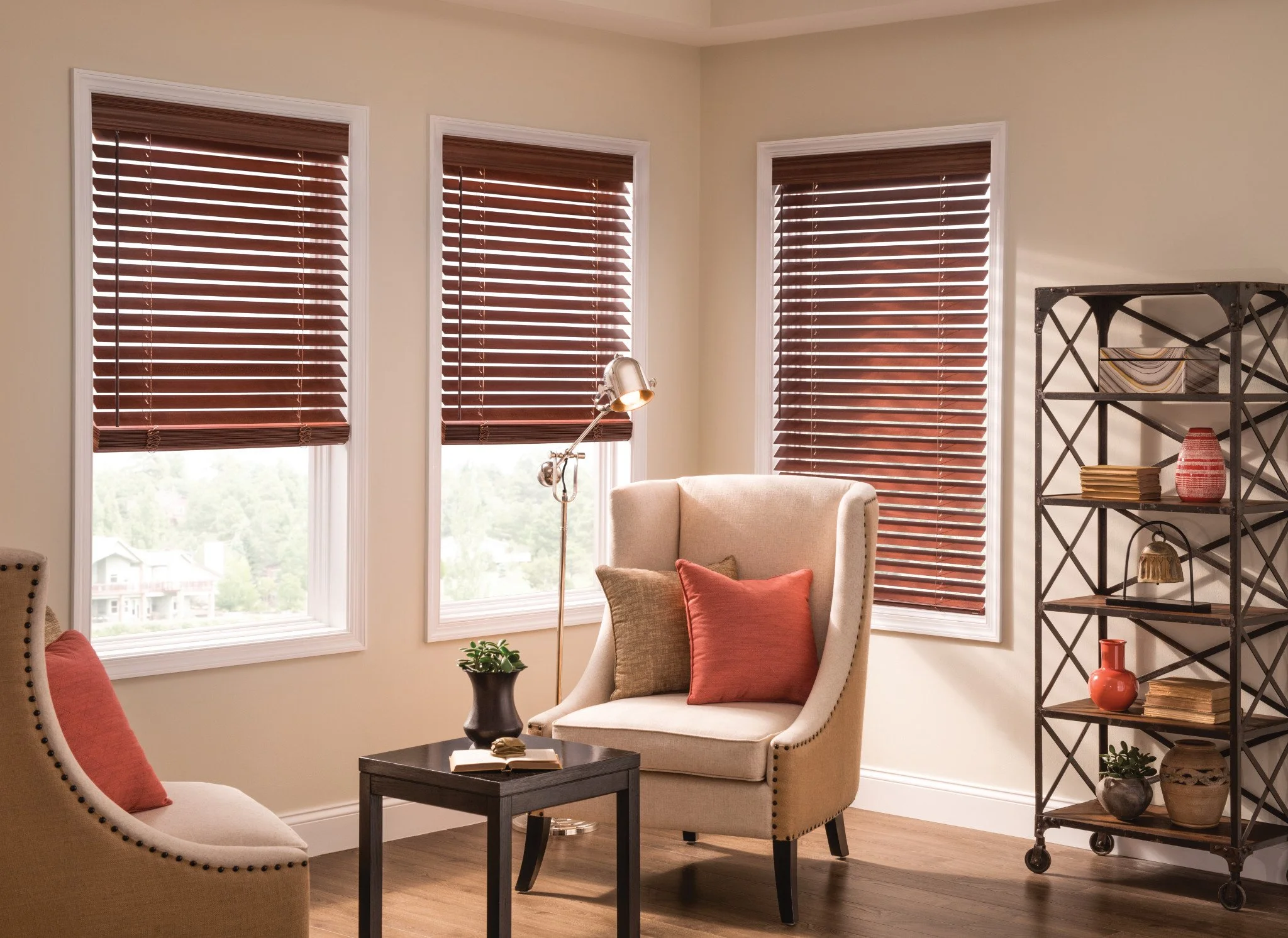 A cozy living room corner with three windows covered by brown wooden blinds, cream-colored walls, beige armchairs with red cushions, a small black side table with a book and a decorative lamp, and an industrial-style black metal bookshelf with decorative items and books.