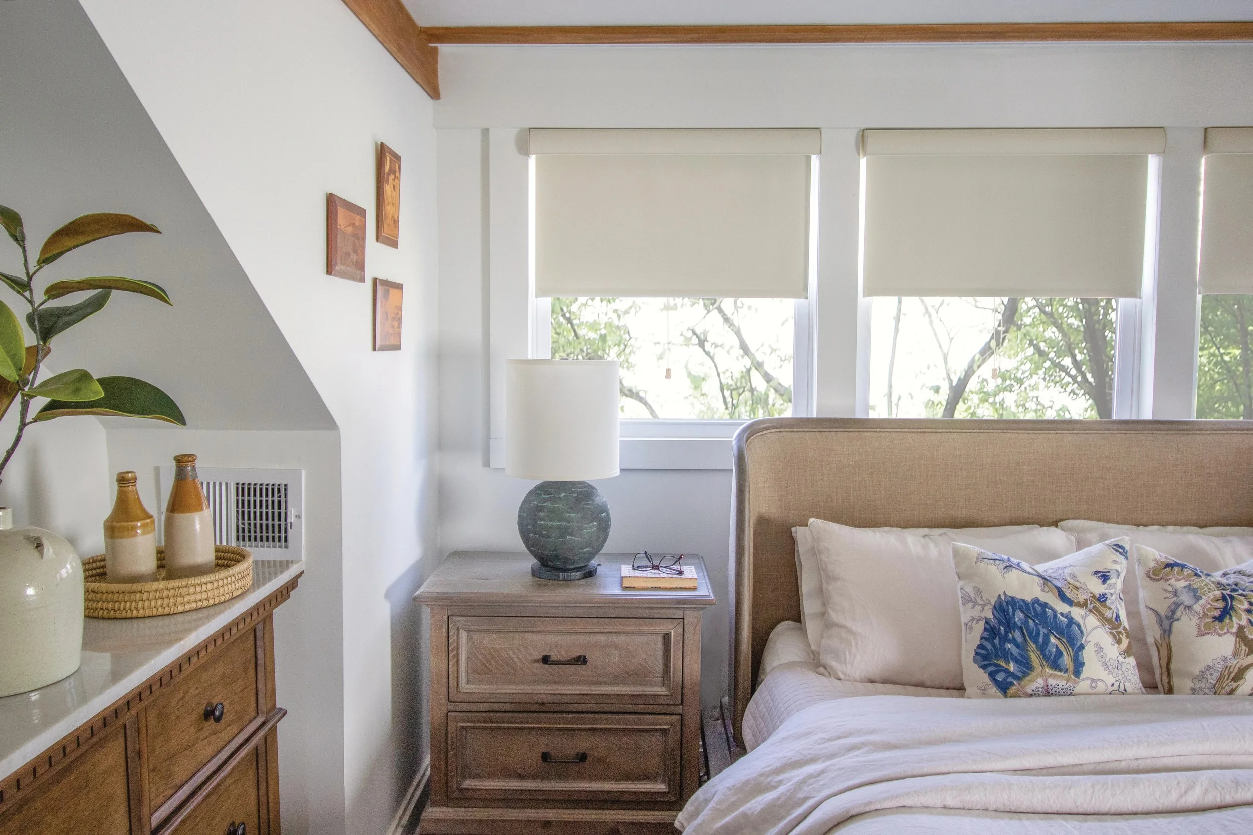 A bedroom with a wooden side table beside the bed, a table lamp, and decorative items including vases and a basket. There are three framed pictures on the wall and two large windows with roll-up blinds, showing trees outside. Part of a green leafy plant is visible on the left.