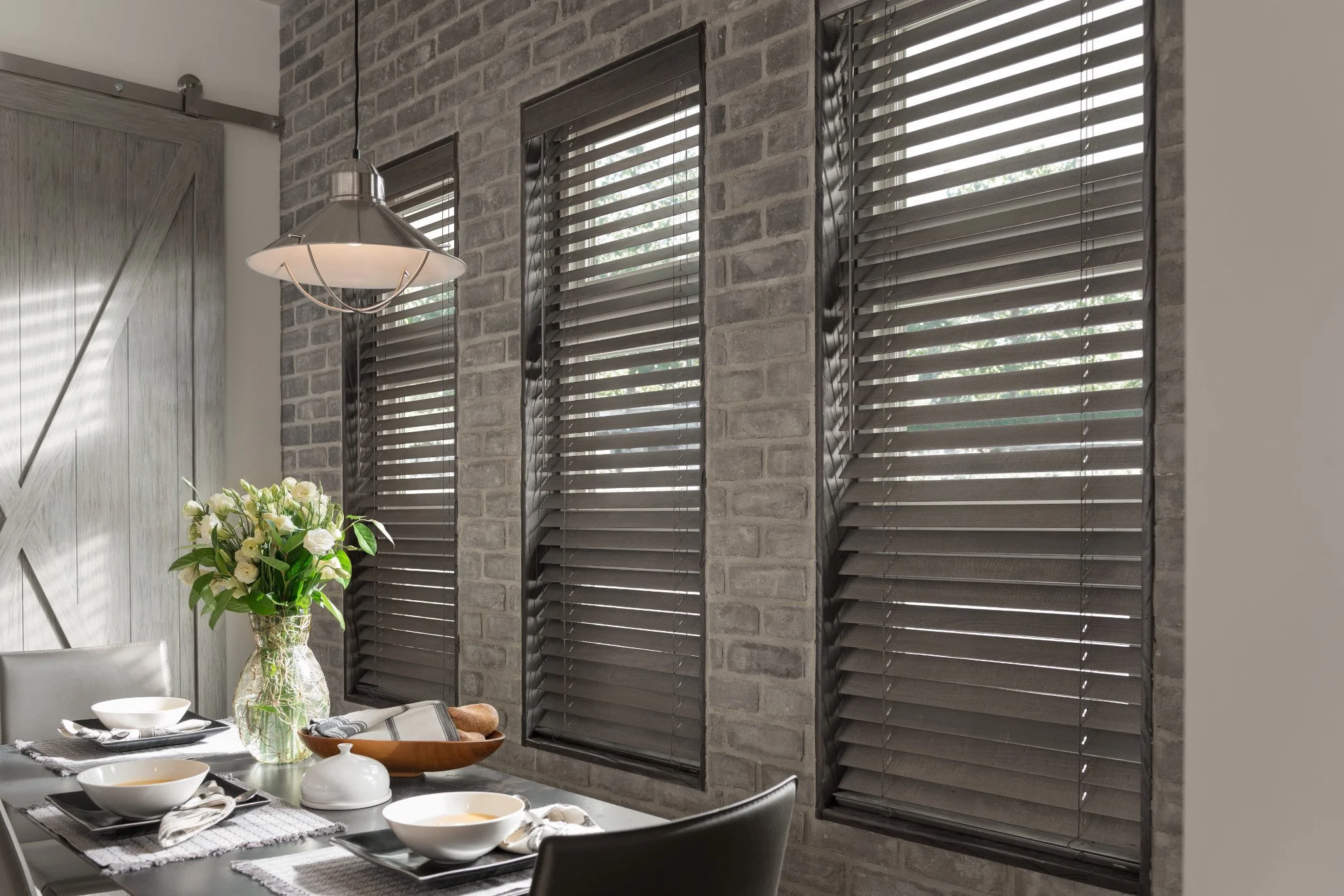 Interior of a dining area with a table set with bowls, plates, and decorative items, a vase with white flowers, and four large windows with dark blinds on a brick wall