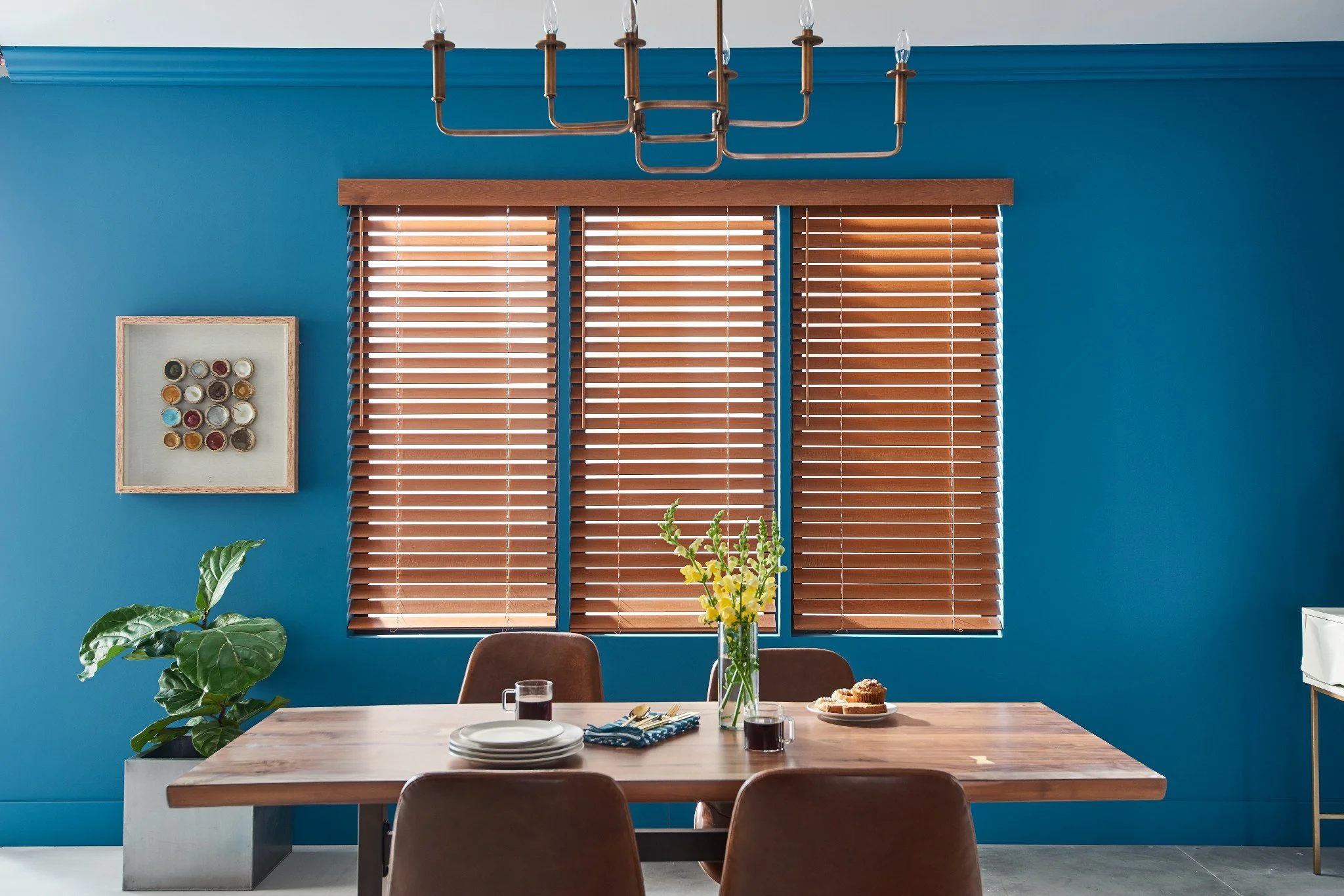 Interior of a dining room with a wooden table set with plates, glasses, and a vase with yellow flowers. The wall is painted bright blue and has wooden blinds covering windows. A framed display of colorful tea tins hangs on the wall, a potted plant is on the left, and a white sideboard is on the right.