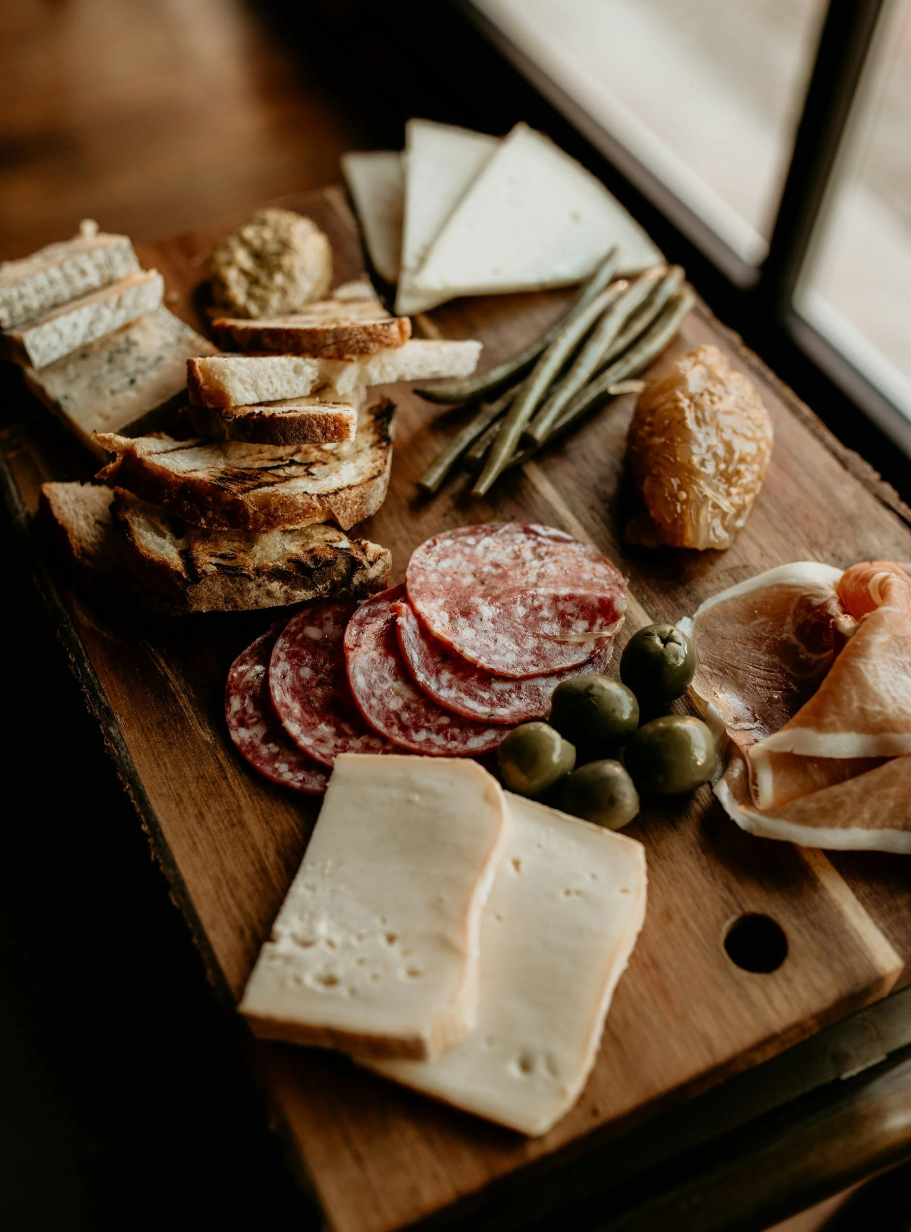 A wooden cutting board with assorted cheeses, cured meats, olives, crackers, and green beans near a window.