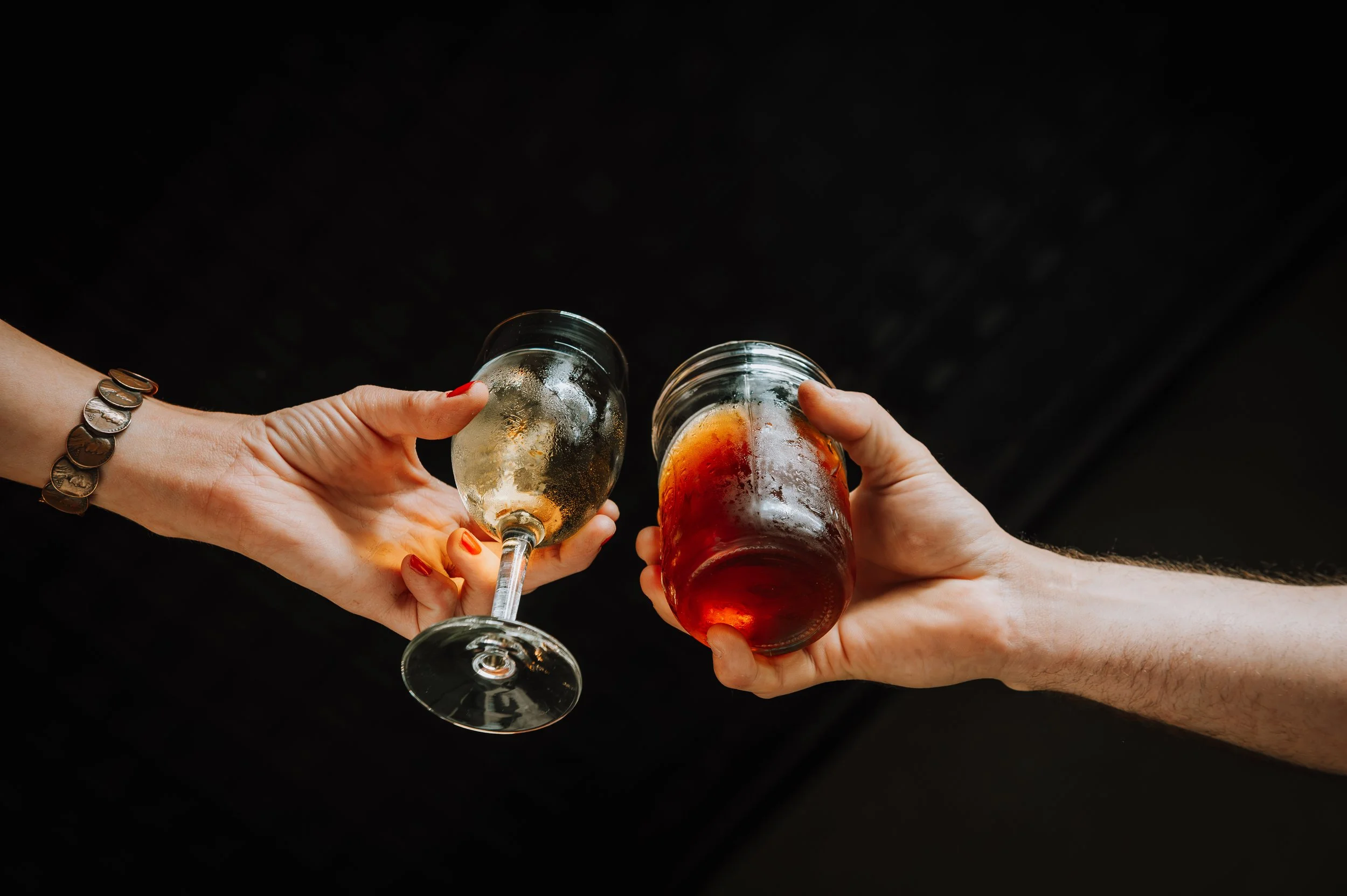 Two people clinking glasses: one with a glass of white wine and the other with a glass of red wine, against a dark background.