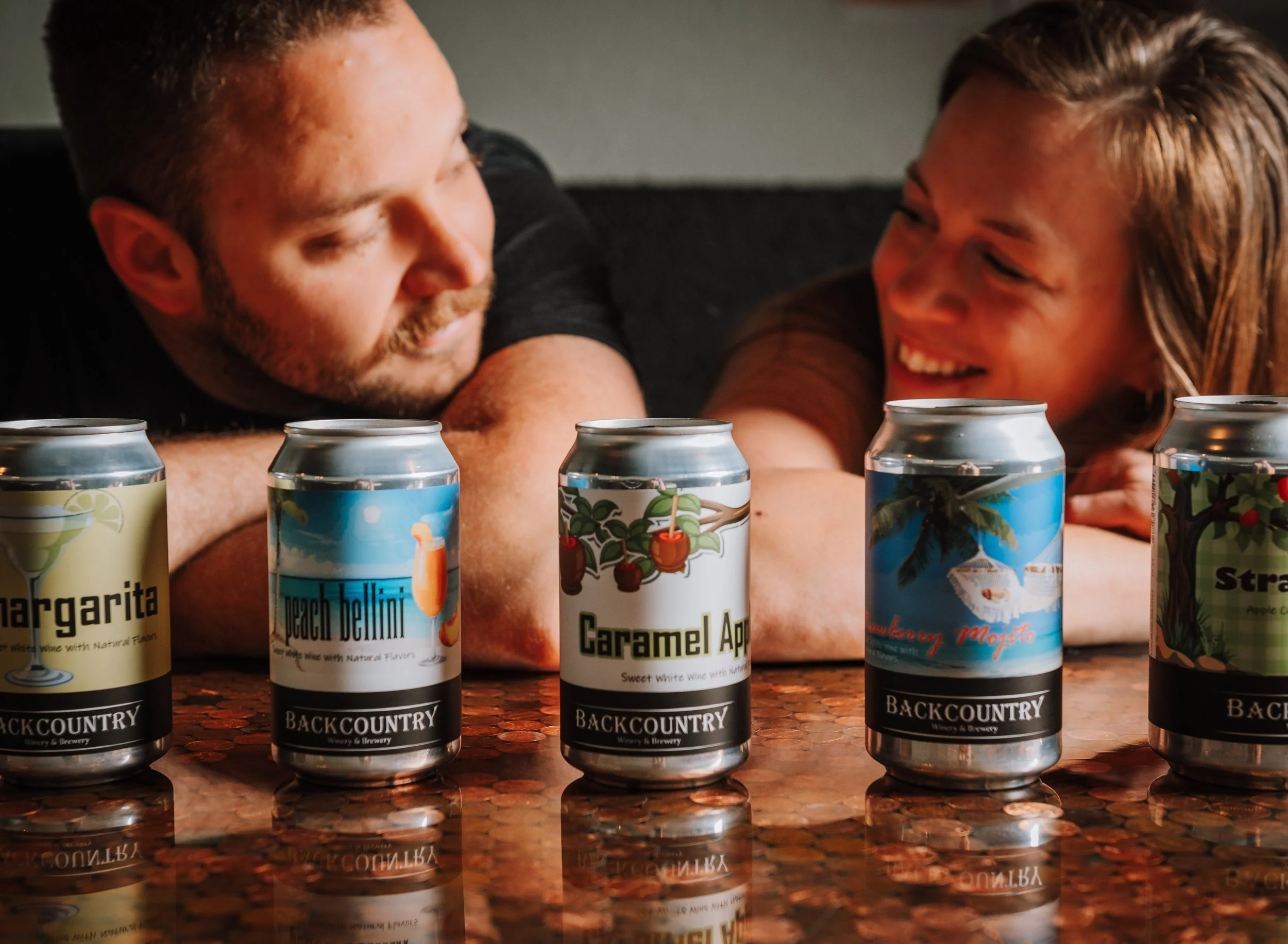 A smiling man and woman leaning on a bar with five cans of flavored wine in between them.