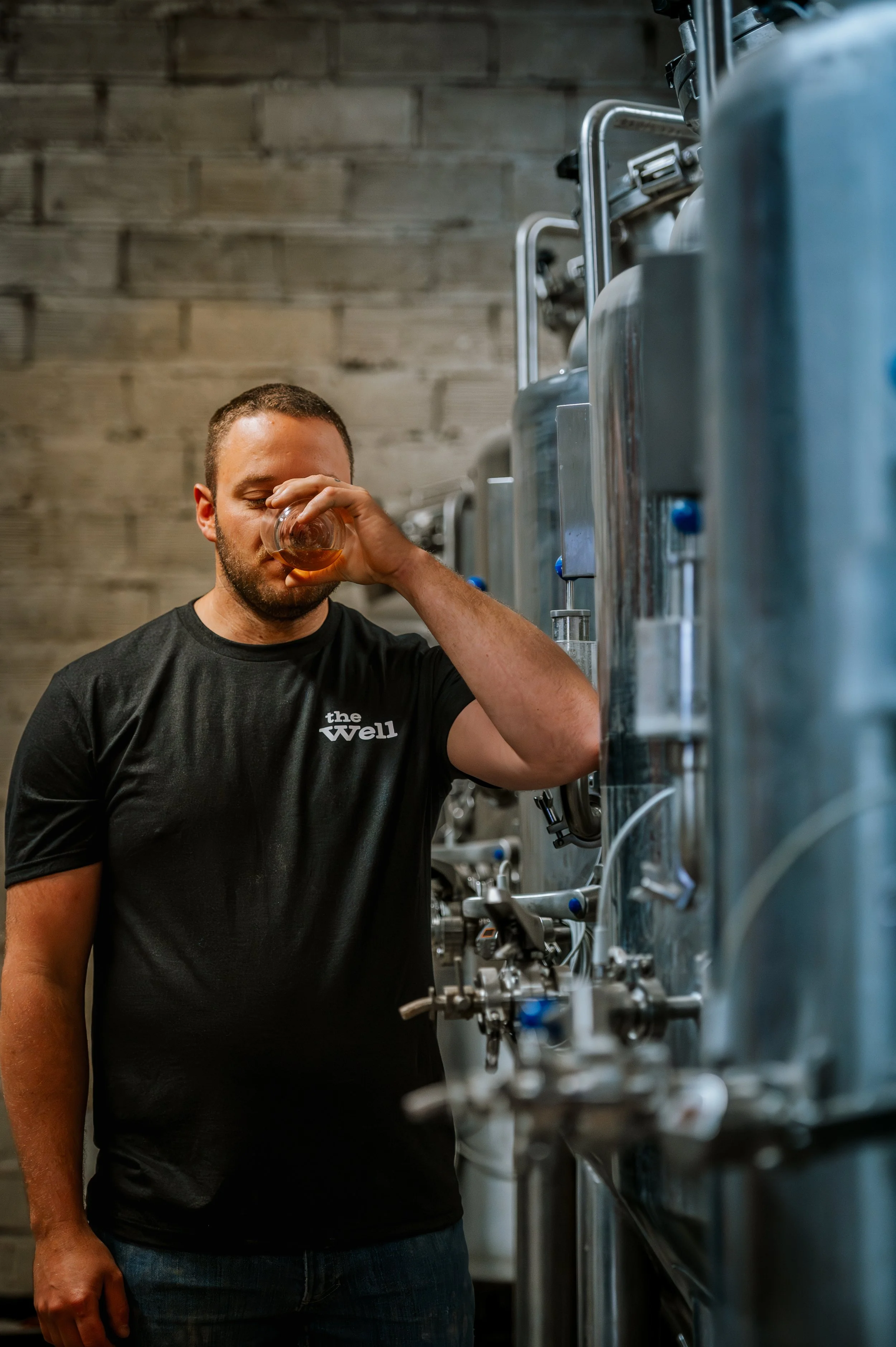 A man in a black shirt is drinking from a glass, standing next to large stainless steel tanks in a brewery.