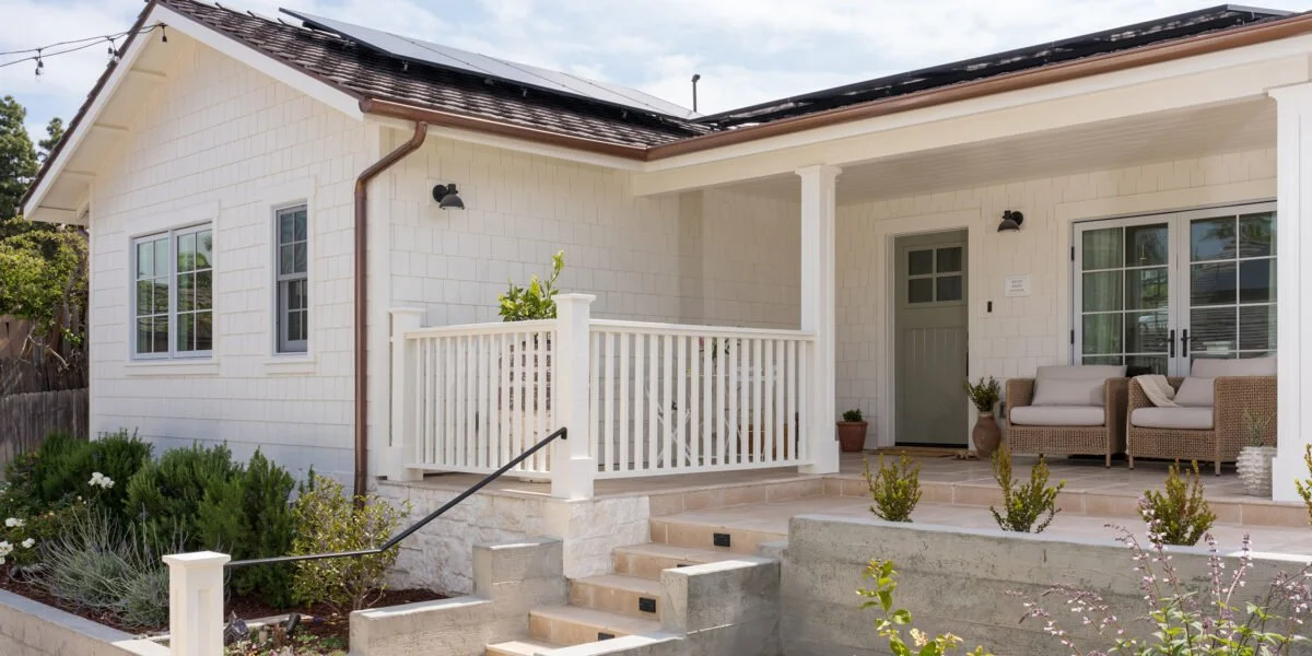 A house with white siding, a front porch with wicker chairs, potted plants, and steps leading up to it, surrounded by greenery.