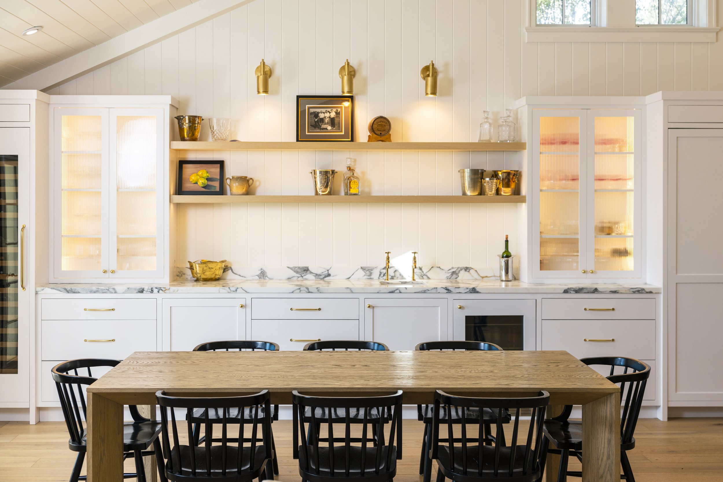 Modern kitchen with white cabinetry, open wooden shelves, marble backsplash, and a wooden dining table with black chairs.
