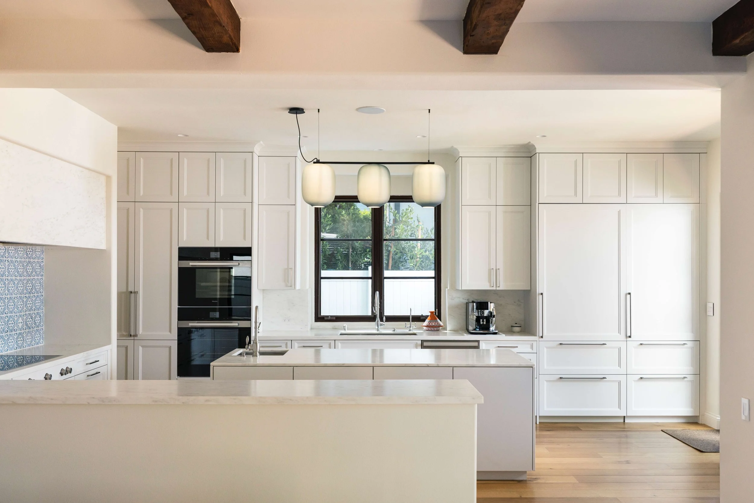 Modern kitchen with white cabinets, a large window, a black oven, and a kitchen island with a marble countertop. Three pendant lights hang above the island, and there is a coffee maker on the counter.