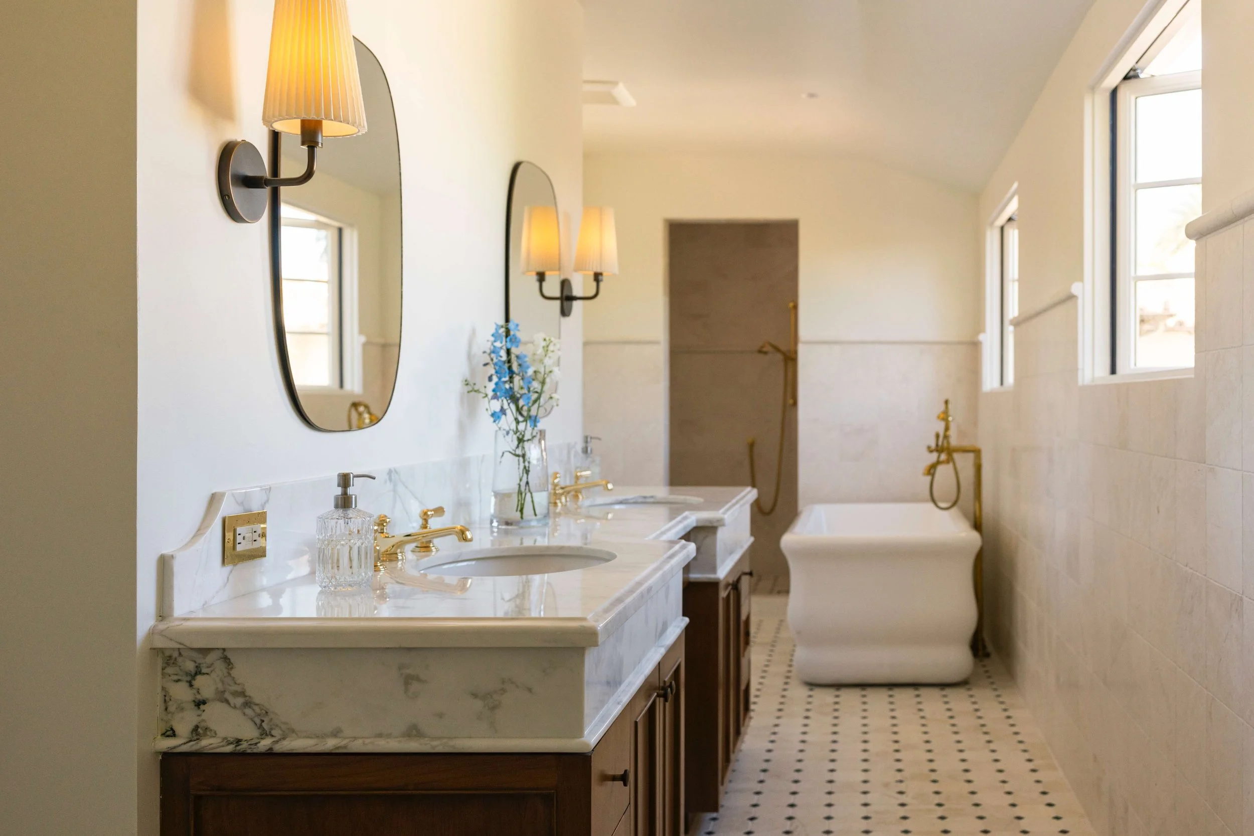 Bright bathroom with double marble sinks, gold fixtures, mirrors, wall sconces, and a vintage bathtub near windows.