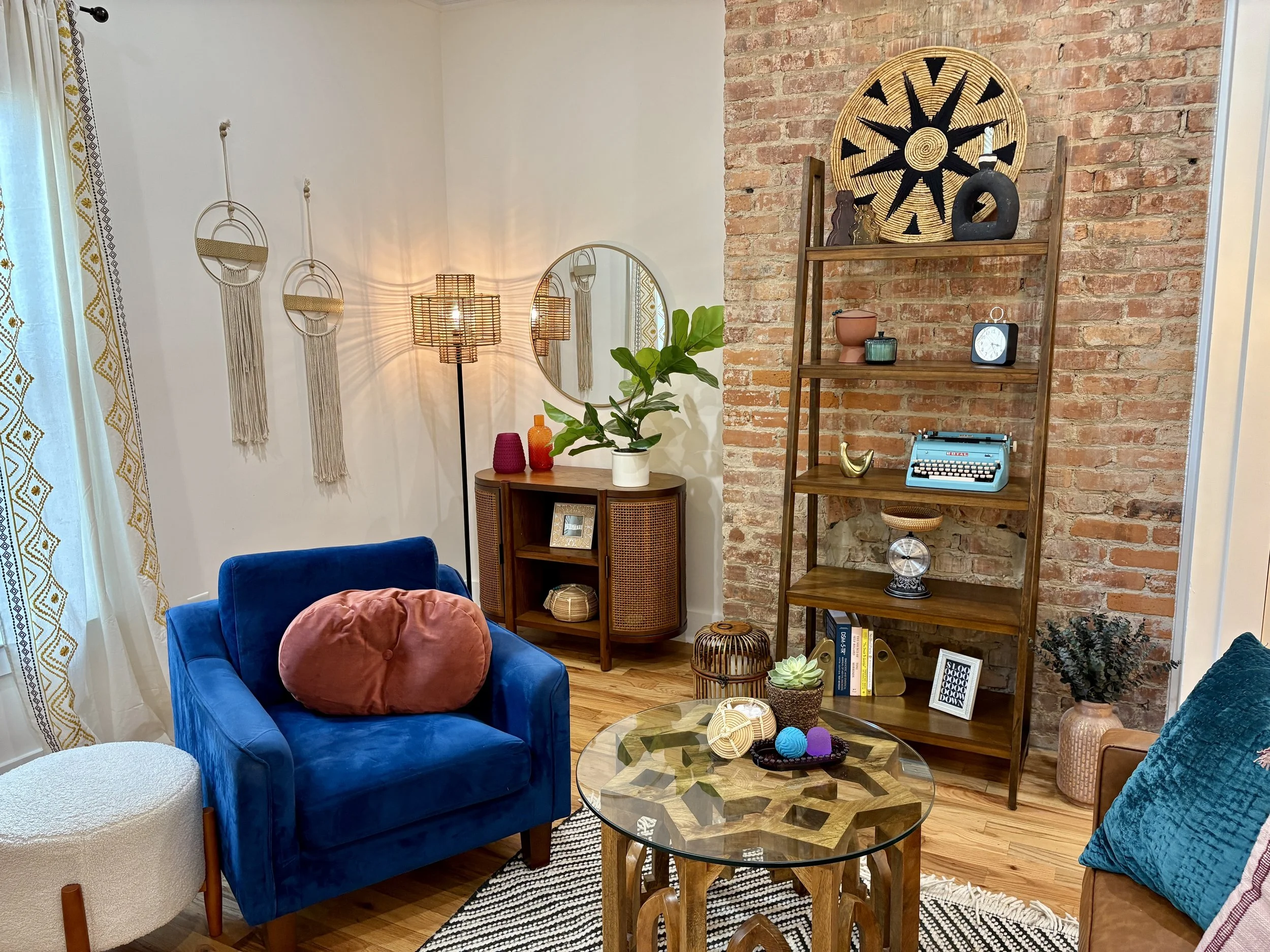 A cozy living room with a brick accent wall, wooden shelving filled with decorative items, a blue velvet armchair with a rust-colored pillow, and a round glass coffee table with colorful decor. There is a green plant on a small cabinet, a standing floor lamp, and hanging wall decor. Light filters in through a window with white curtains.