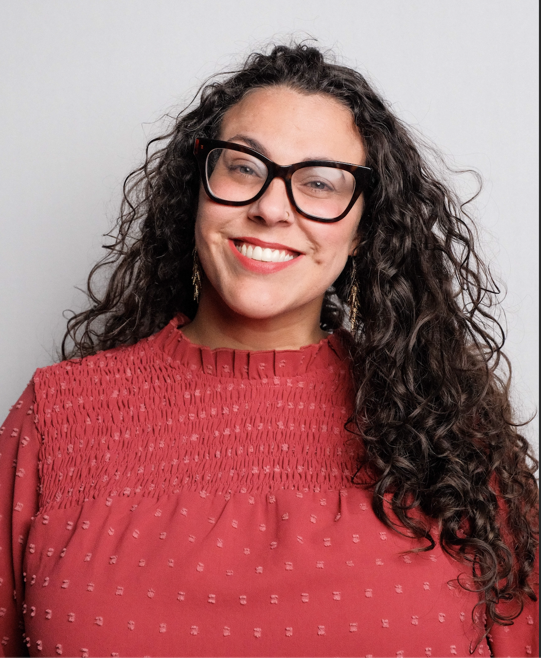 A woman with curly hair, wearing glasses and earrings, smiling, in a red blouse with textured detailing and a high neckline, standing against a plain white background.