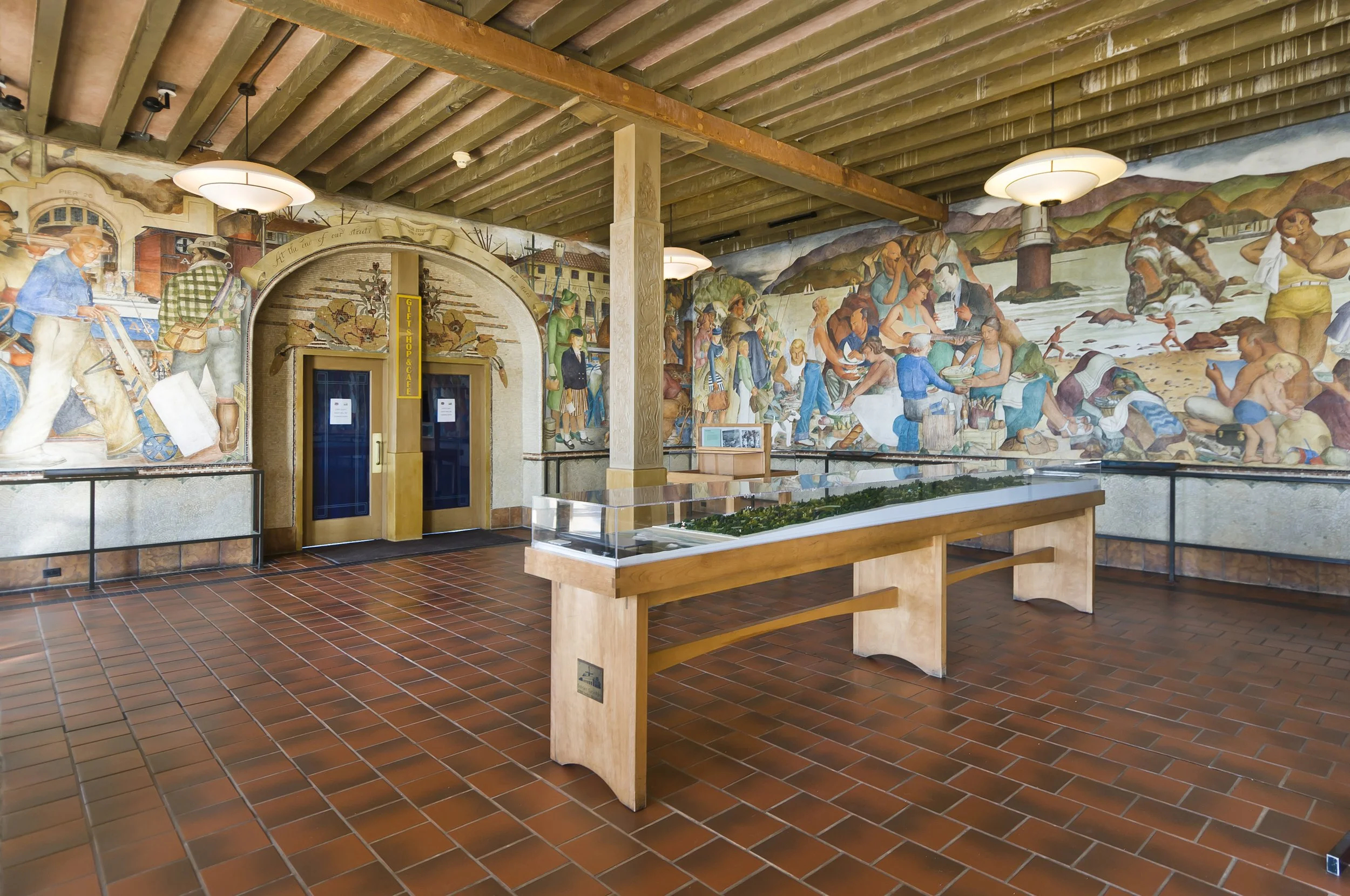 Indoor space with mural wall depicting historical scenes, a model display case, tiled floor, double glass doors, and ceiling lights.