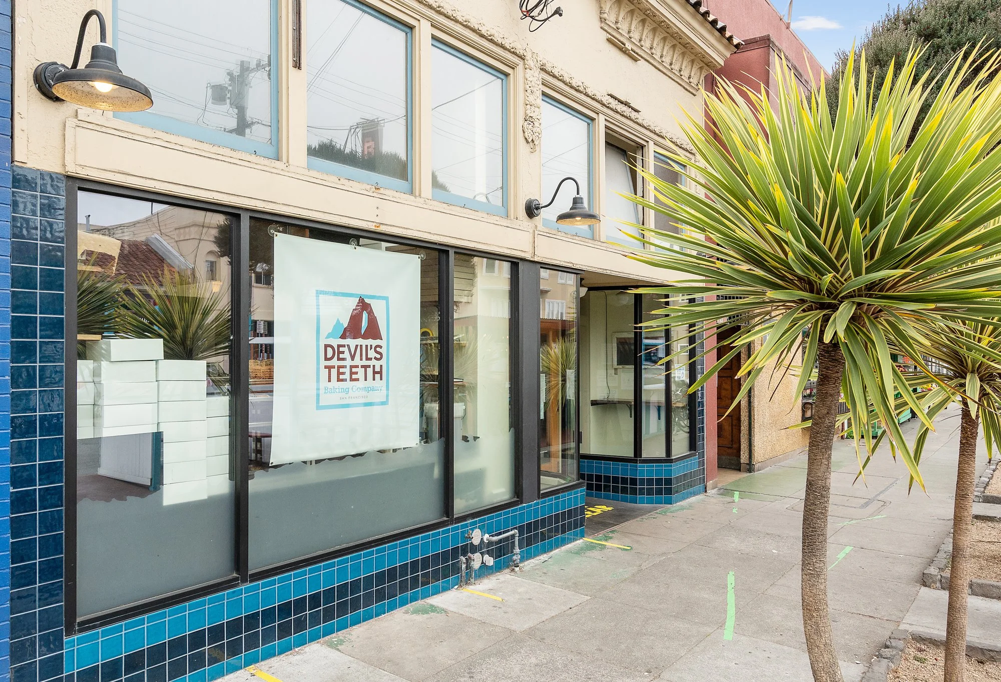 Front view of a building with blue tile accents, large glass windows, and a sign that reads "Devil's Teeth Baking Company, San Francisco." Two trees with long, pointed leaves are on the sidewalk in front.