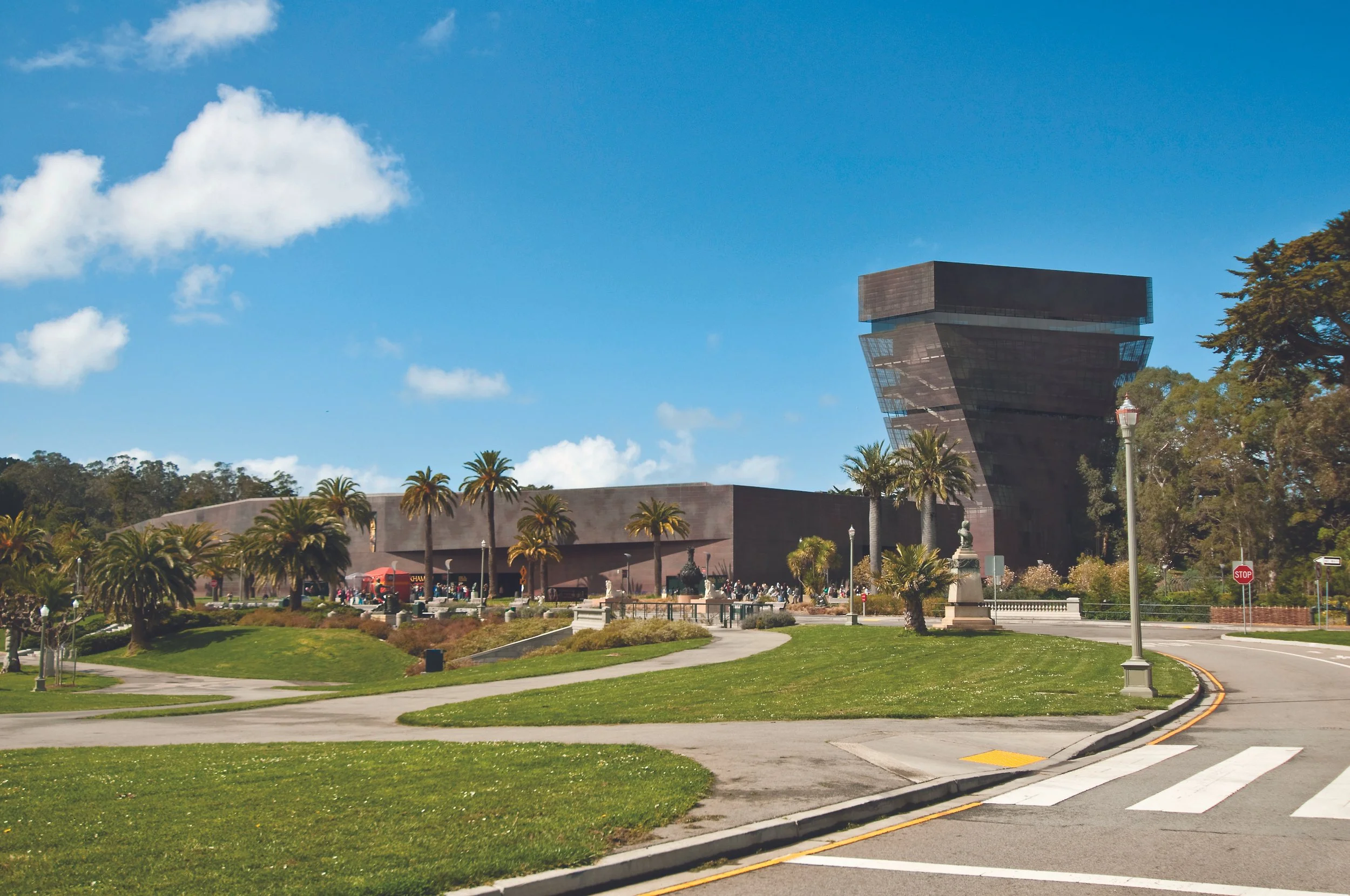 A modern building with a unique, tilted architectural design, surrounded by palm trees, grass, and a pedestrian walkway on a sunny day with a blue sky and some clouds.