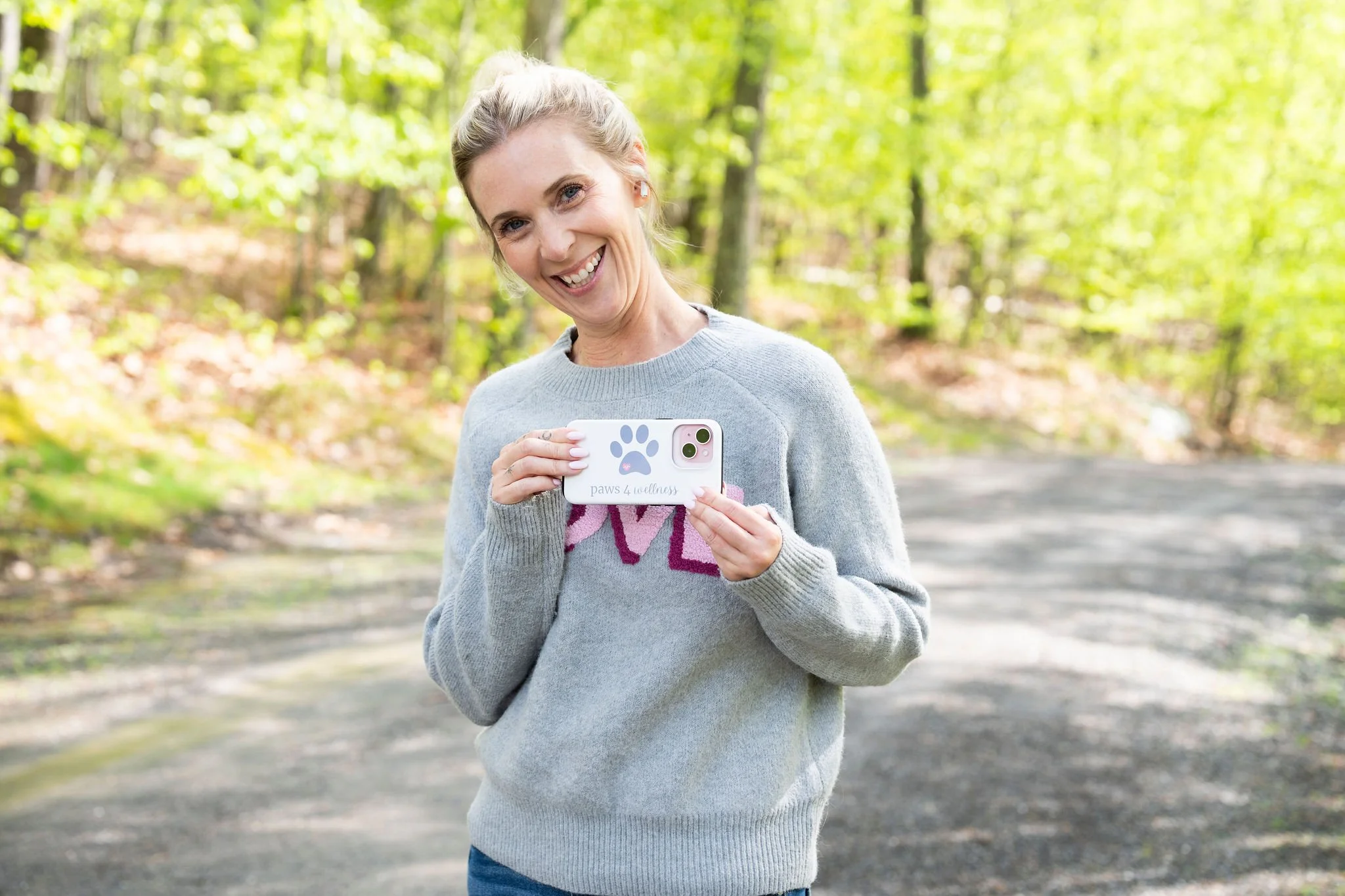 Woman with blonde hair smiling and holding a white phone with paw print design in an outdoor park with green trees.