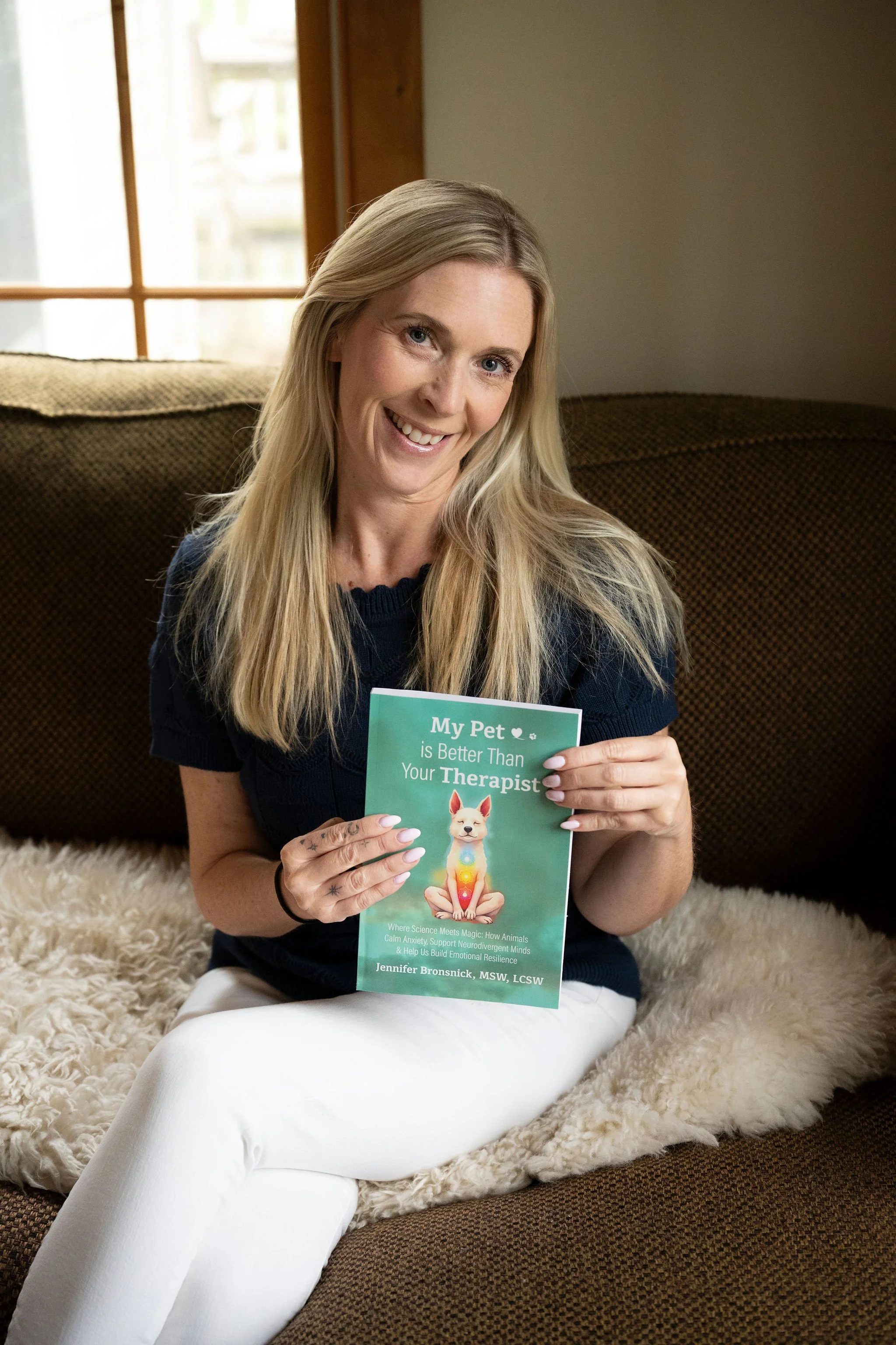 A woman with long blonde hair sitting on a beige furry rug, holding a book titled 'My Pet is Better Than Your Therapist.'