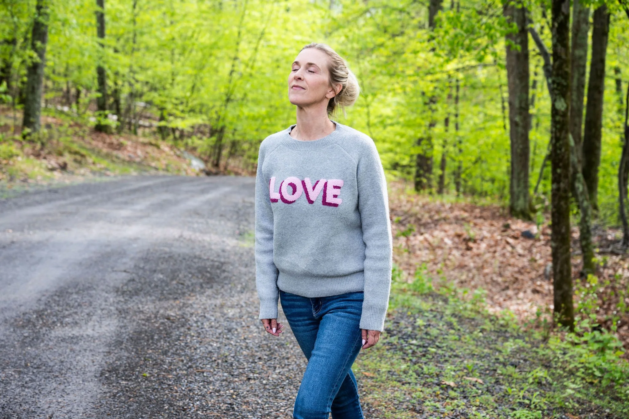 A woman standing on a gravel path in a forest with green leaves, wearing a light gray sweater with the word "LOVE" on it and blue jeans, with her eyes closed and a peaceful expression.