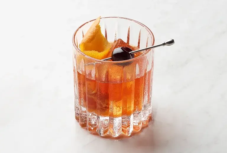 A glass of whiskey on the rocks garnished with a lemon twist and a cherry, with a cocktail pick resting on the glass rim.
